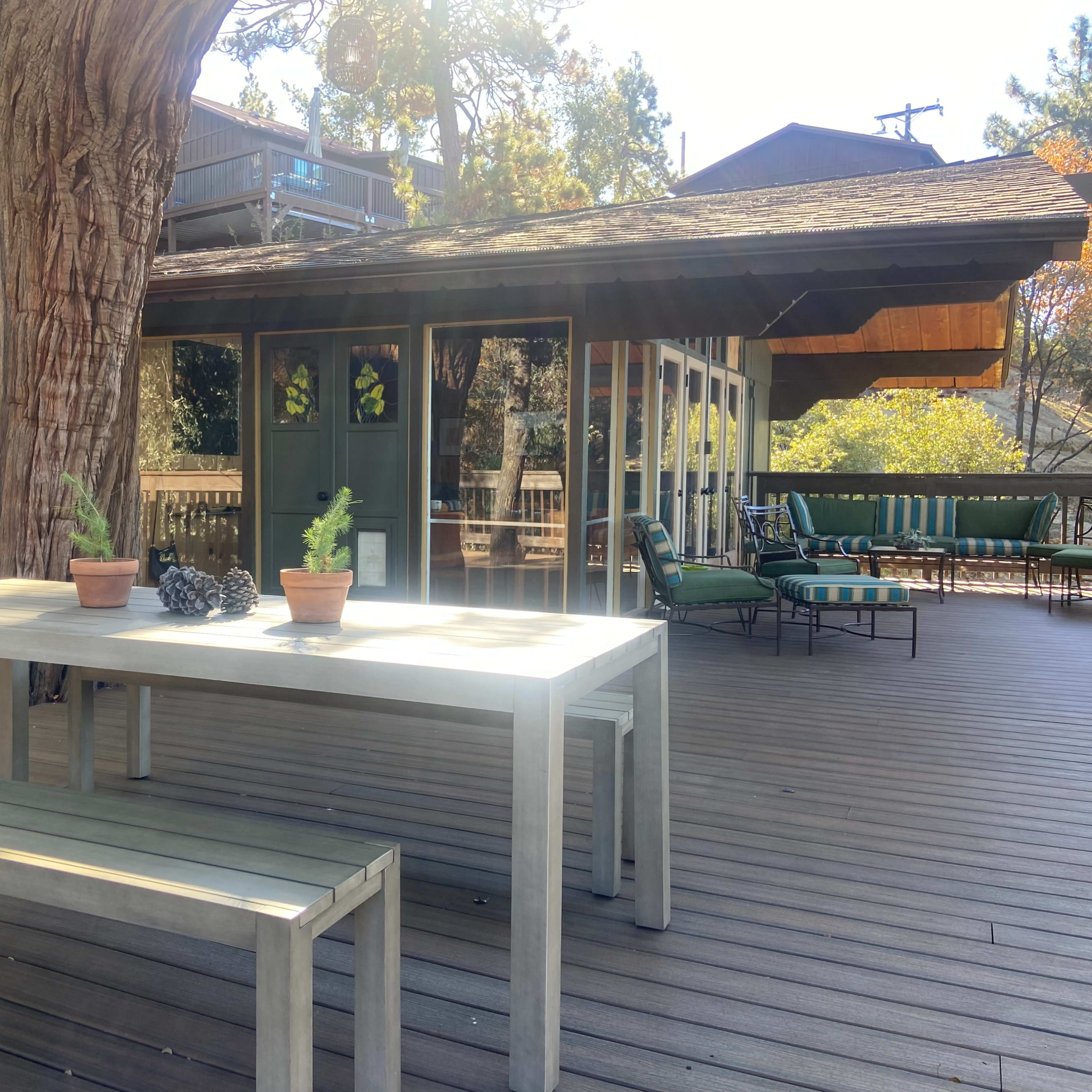 A wooden table with potted plants stands on a deck next to a large tree, overlooking a home with large windows and outdoor seating.