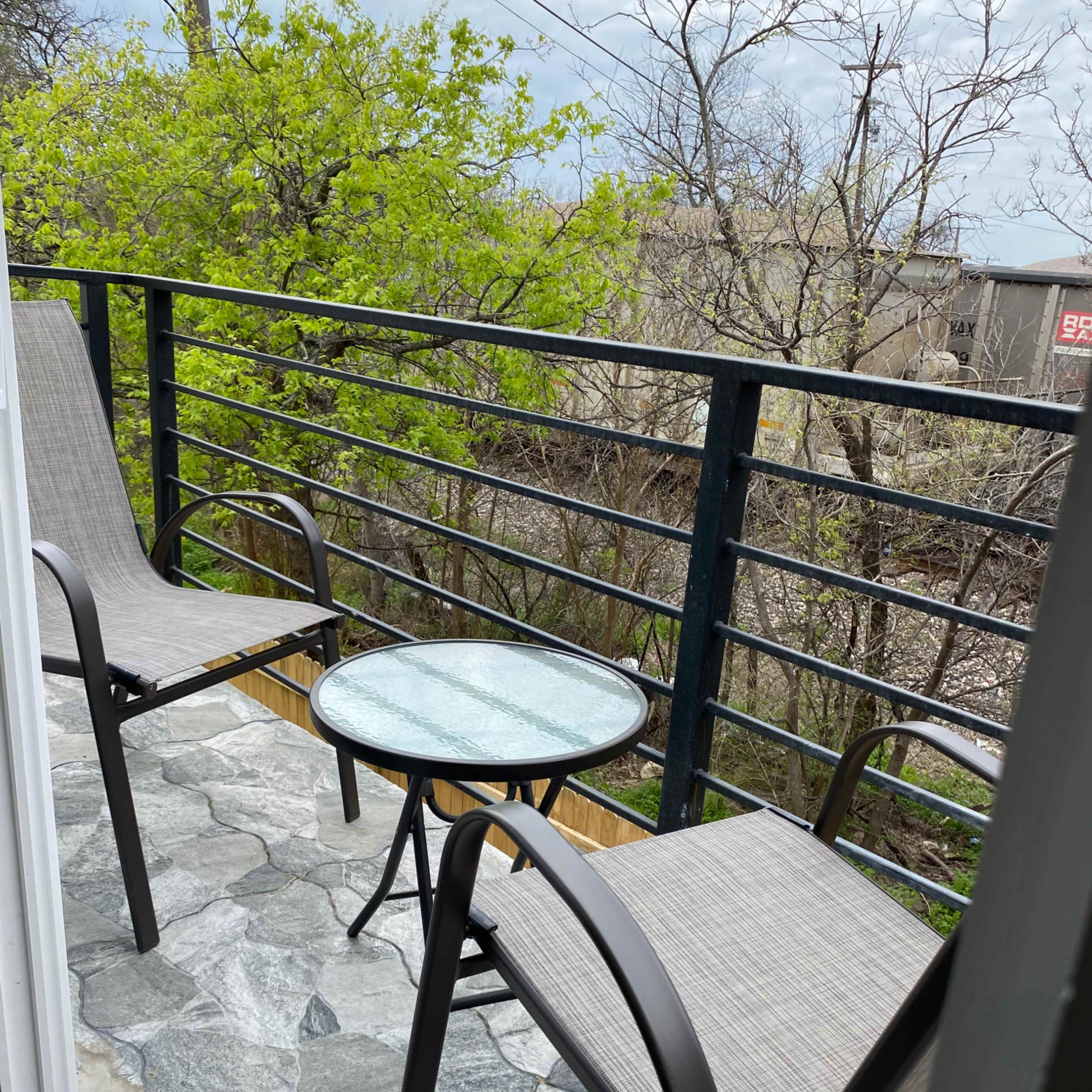 A small balcony with two chairs and a round table, surrounded by greenery and trees.