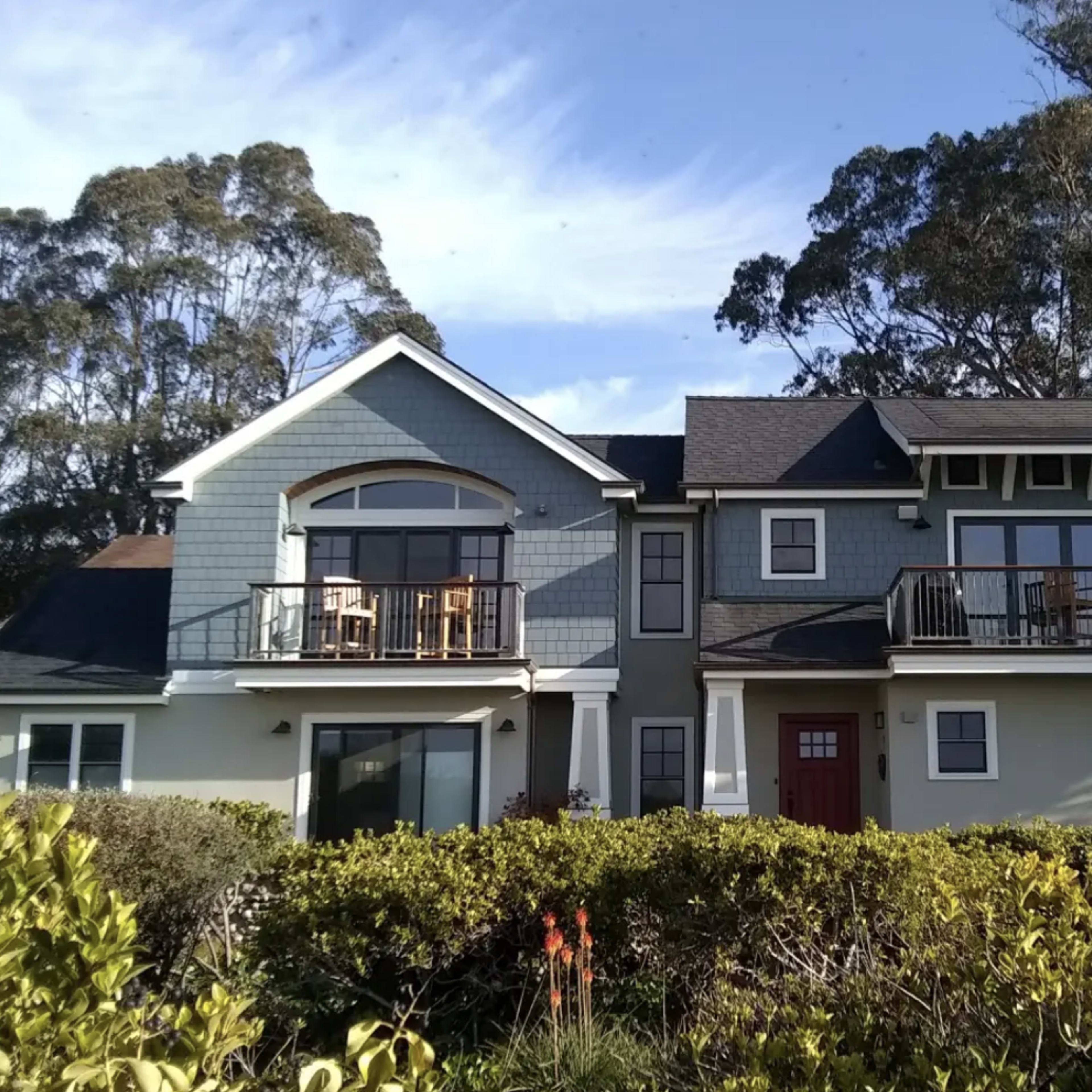 A two-story blue house with balconies and a red front door, surrounded by green shrubs and trees.