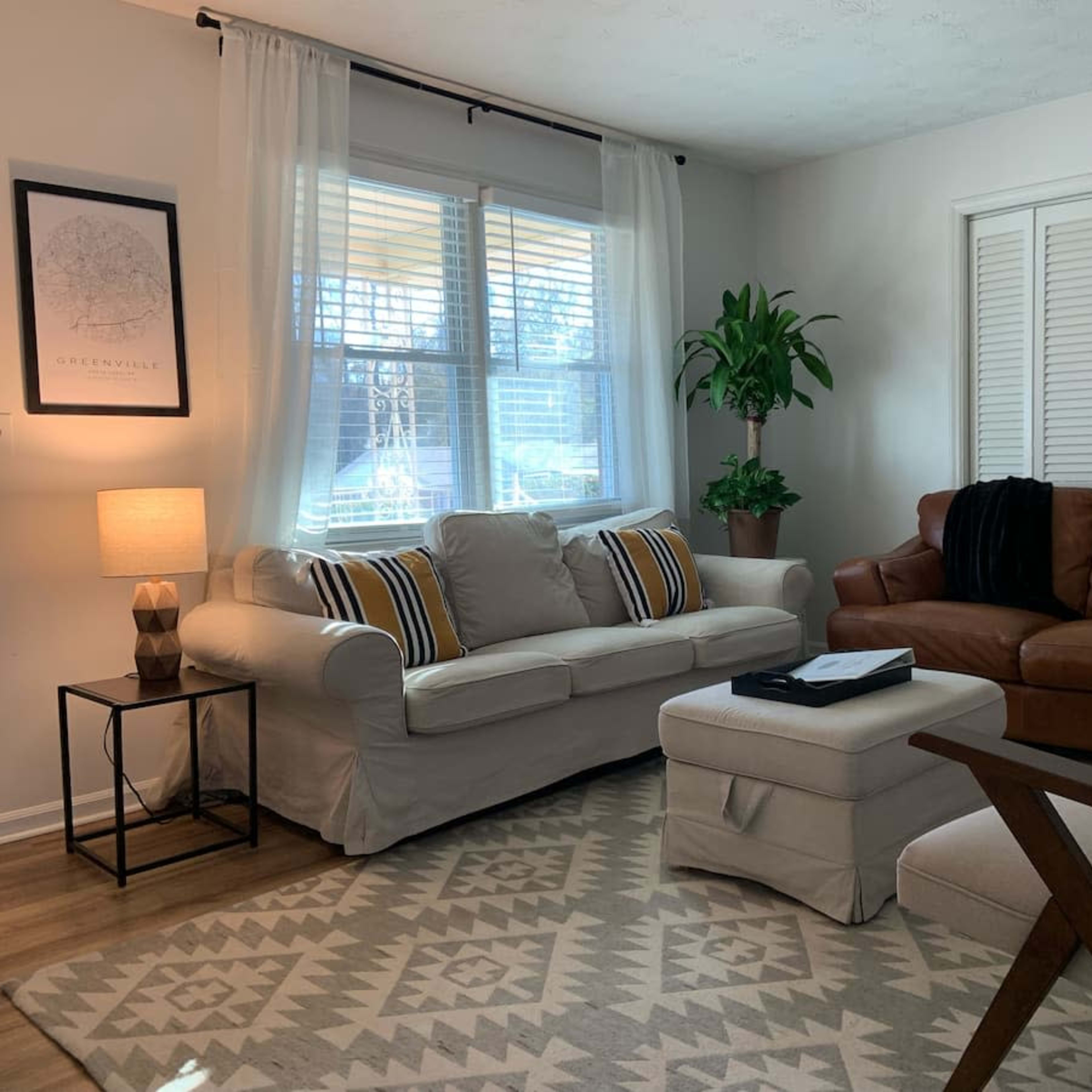 The living room features a light-colored couch and a brown leather sofa, with a small side table and a plant placed near the window.