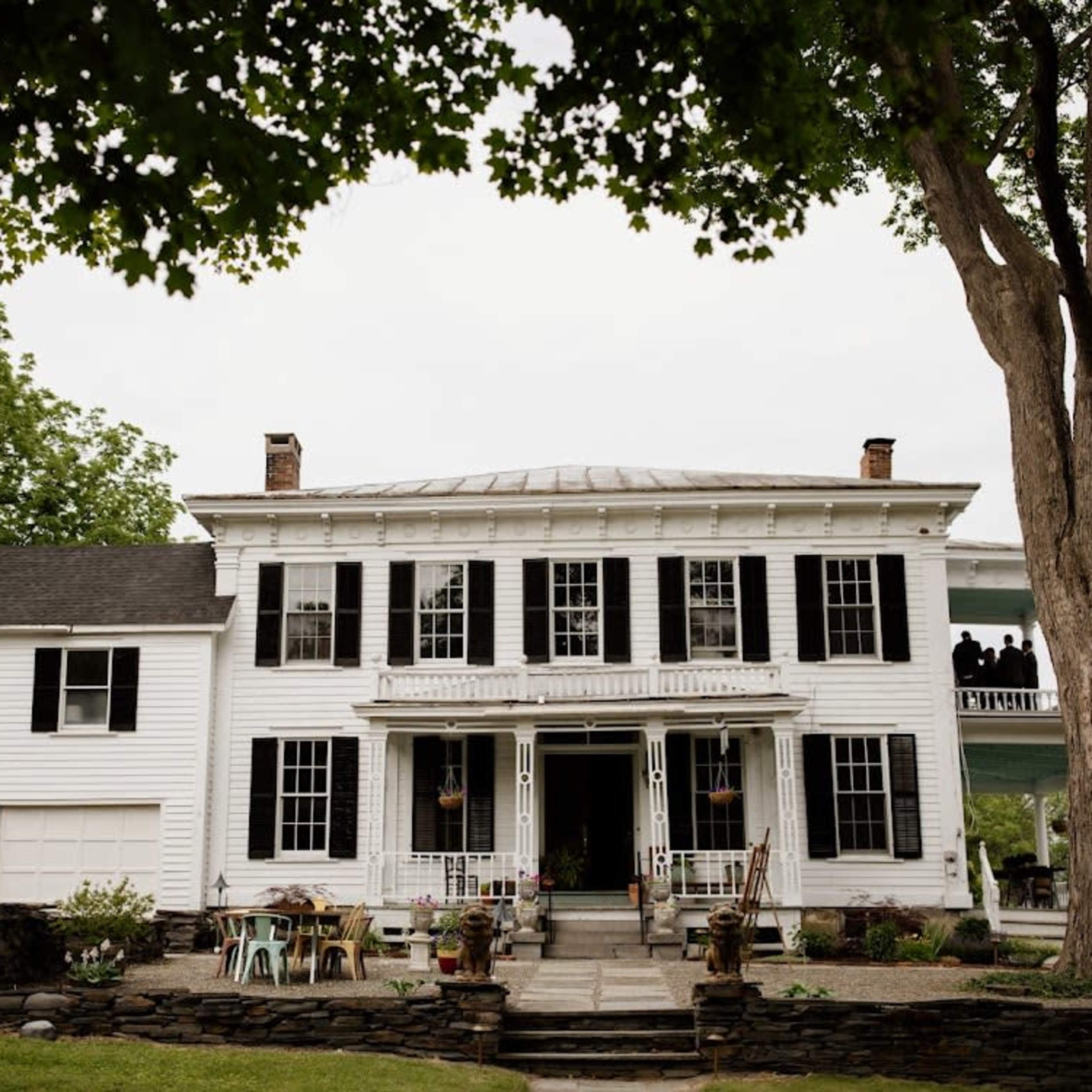A large white house with black shutters, a central entrance, and a porch, surrounded by trees and patio furniture.