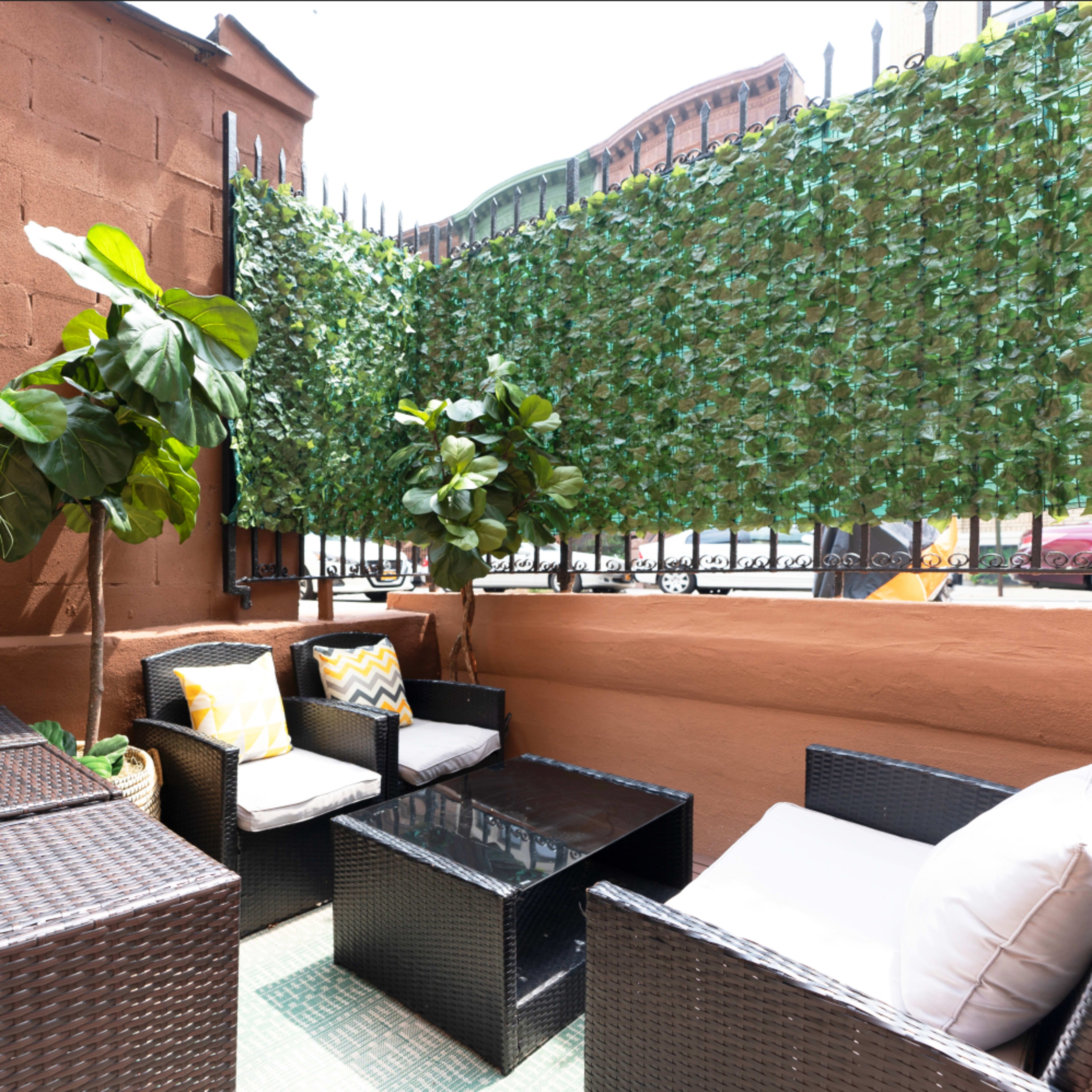 A small outdoor patio area with wicker furniture, potted plants, and a green leafy privacy screen.