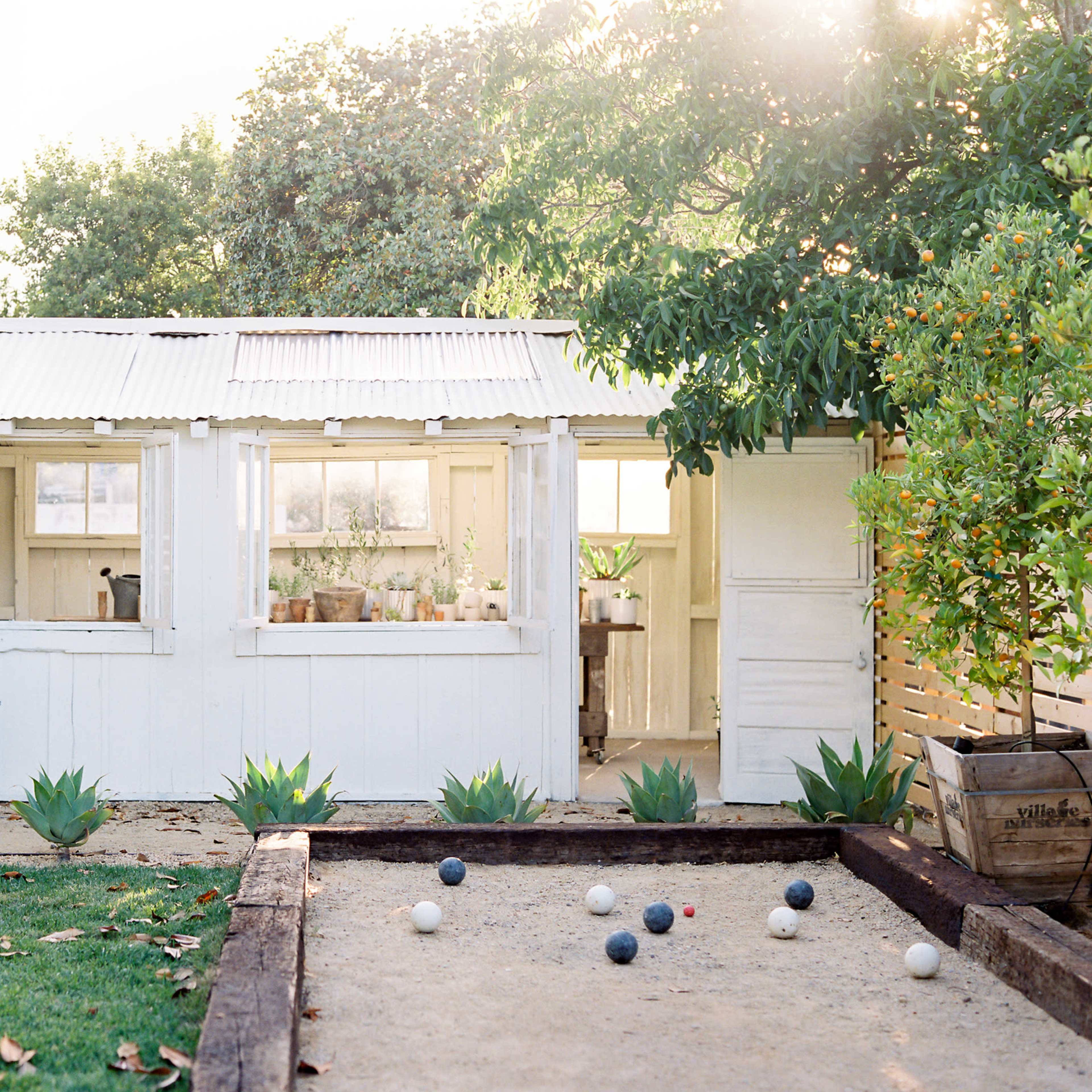 A white shed with large windows stands in a garden, next to a sand court with several balls arranged.