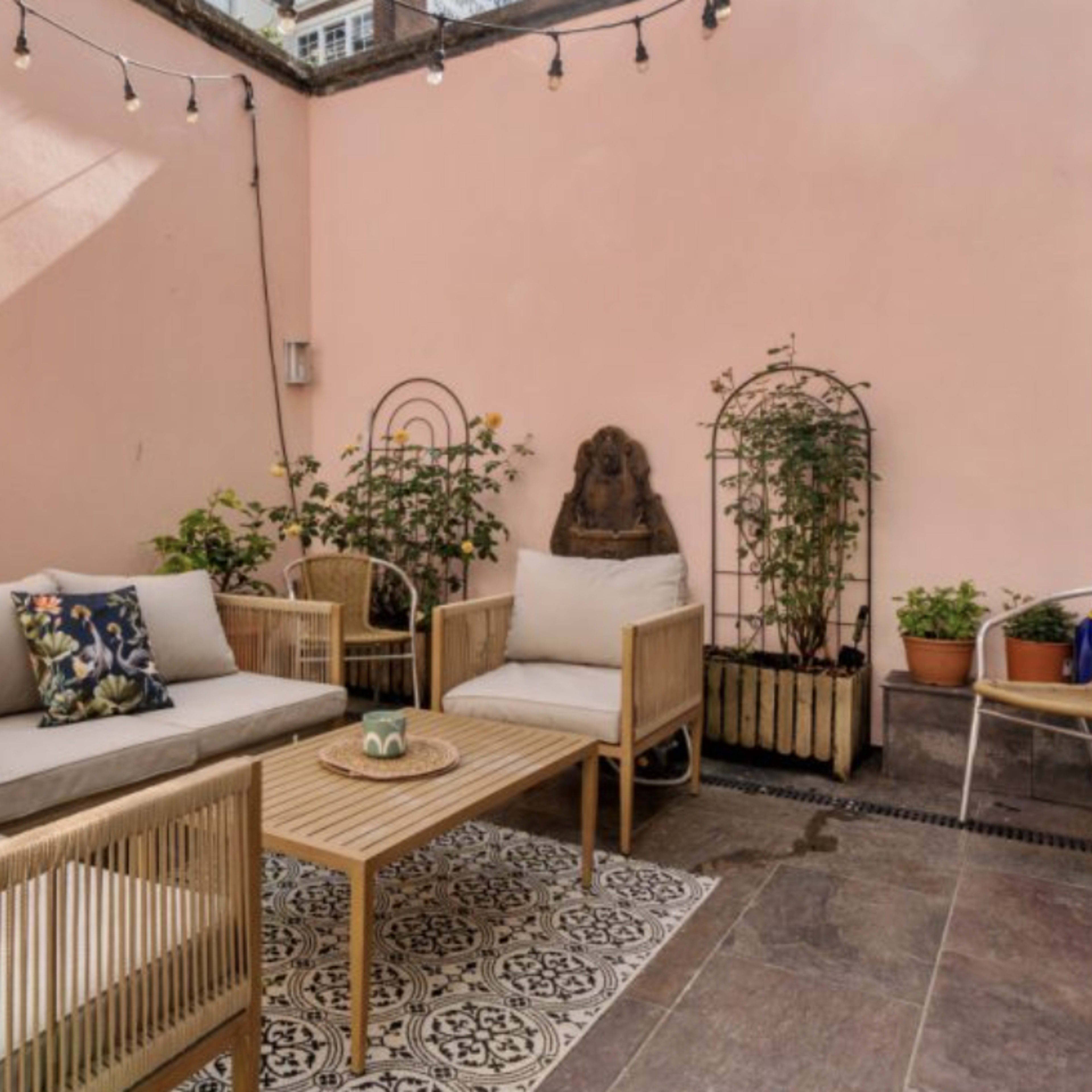 A small outdoor patio features a seating area with wicker furniture, a central wooden coffee table, and potted plants against a light pink wall.