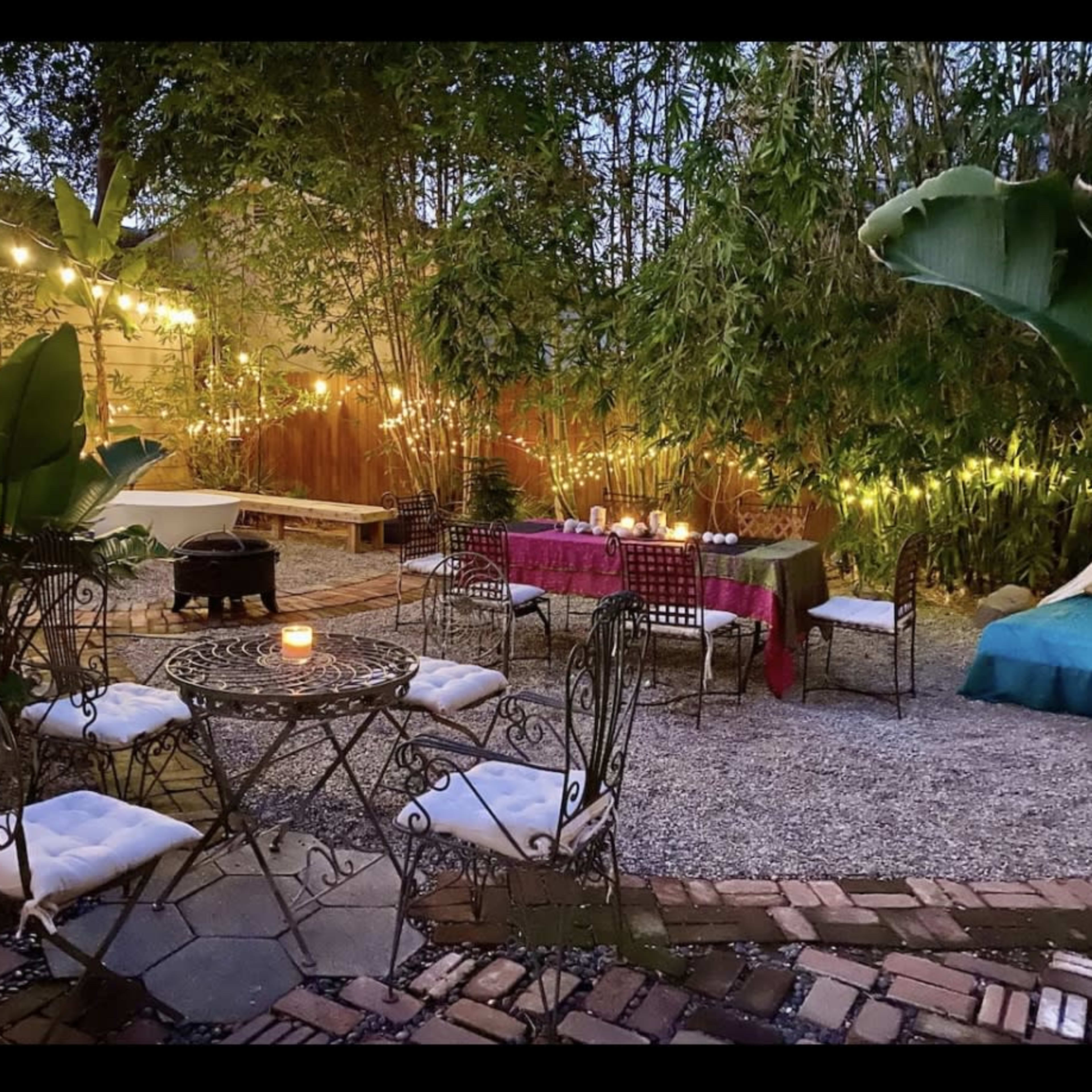 A cozy outdoor seating area features a wrought-iron table surrounded by chairs, illuminated by string lights and complemented by potted plants and a gravel path.