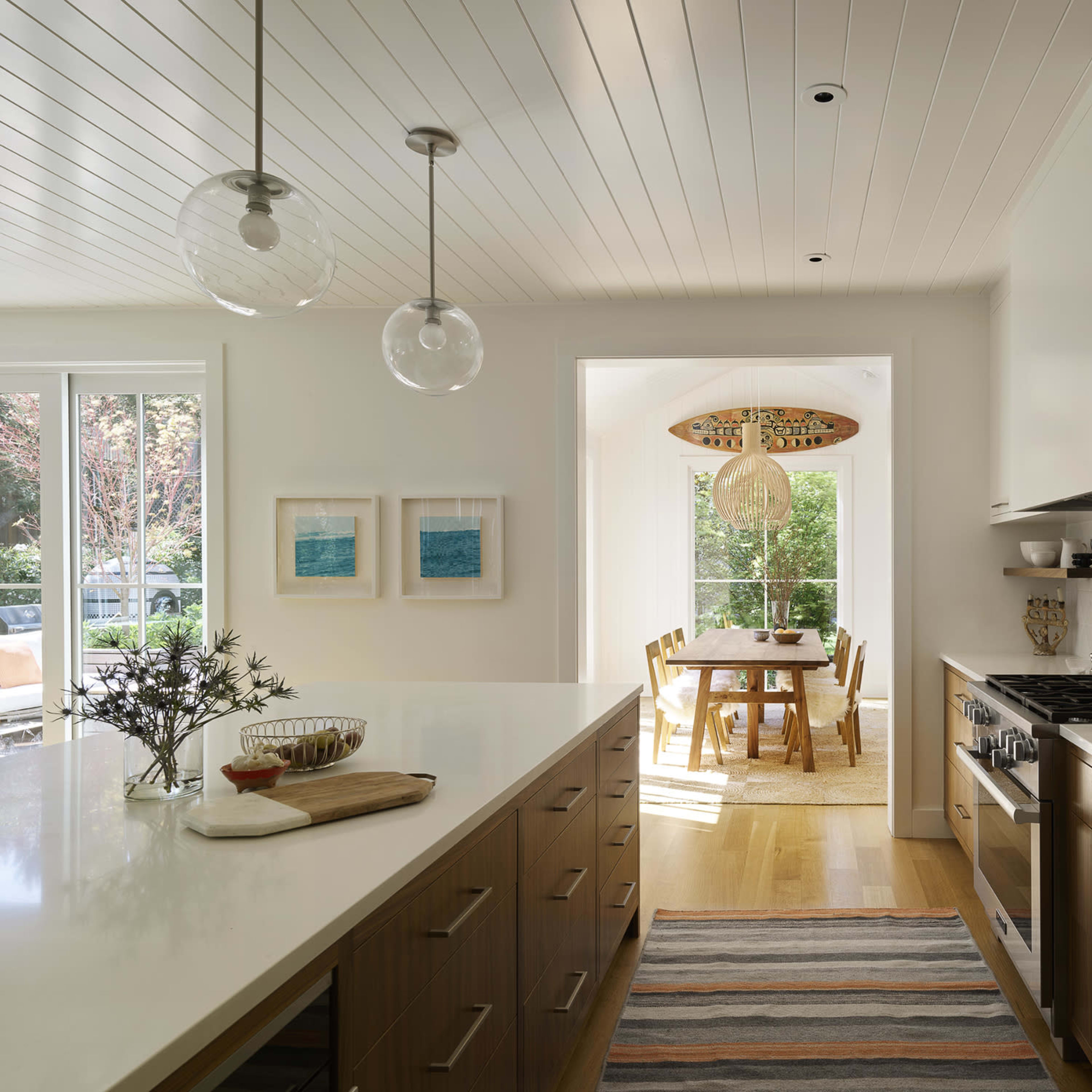 A modern kitchen featuring a large island with a gas stove, light wood cabinetry, and a view into a dining area with a table and chairs, illuminated by natural light.