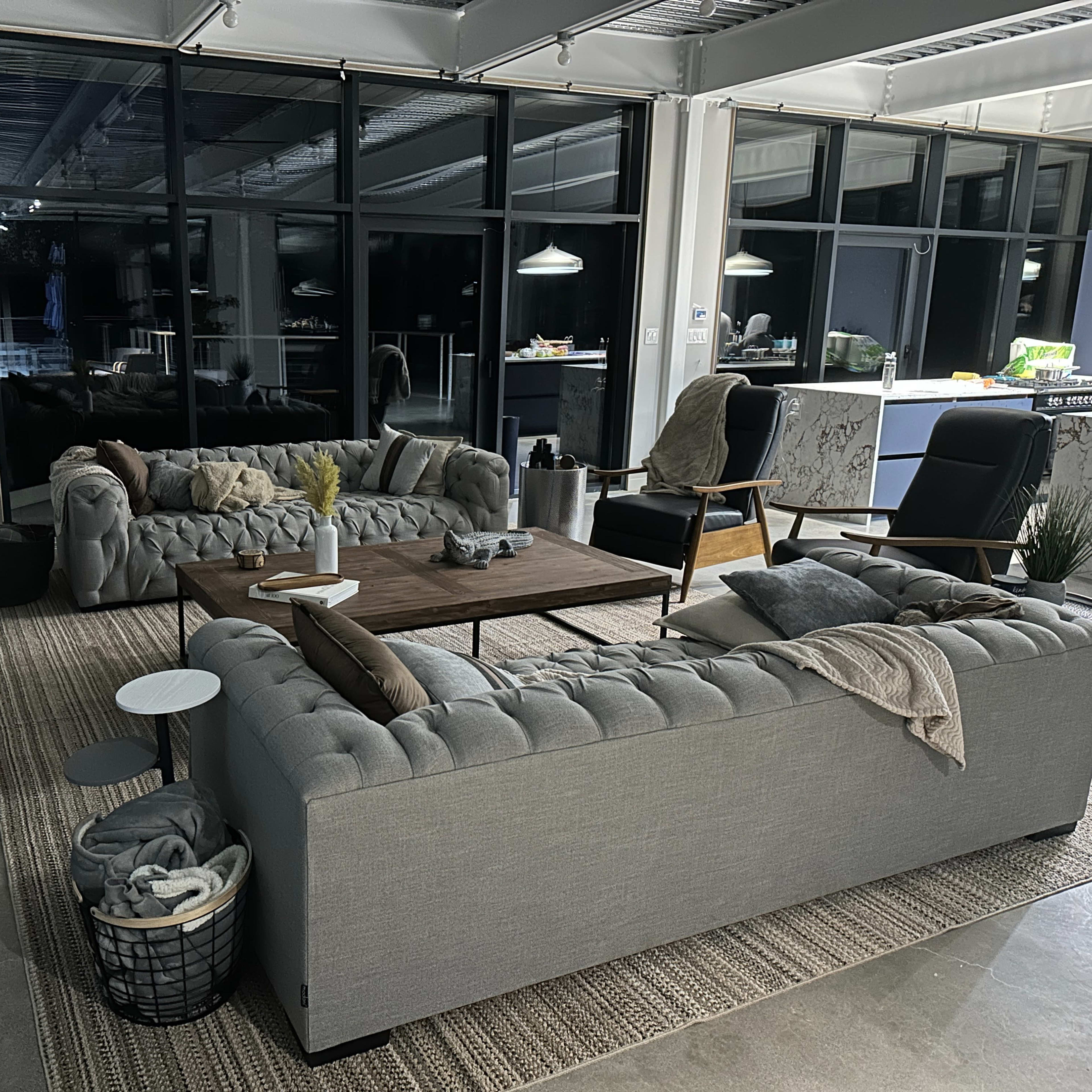 A modern living room with two grey tufted sofas, a wooden coffee table, and large windows allowing ample light in.