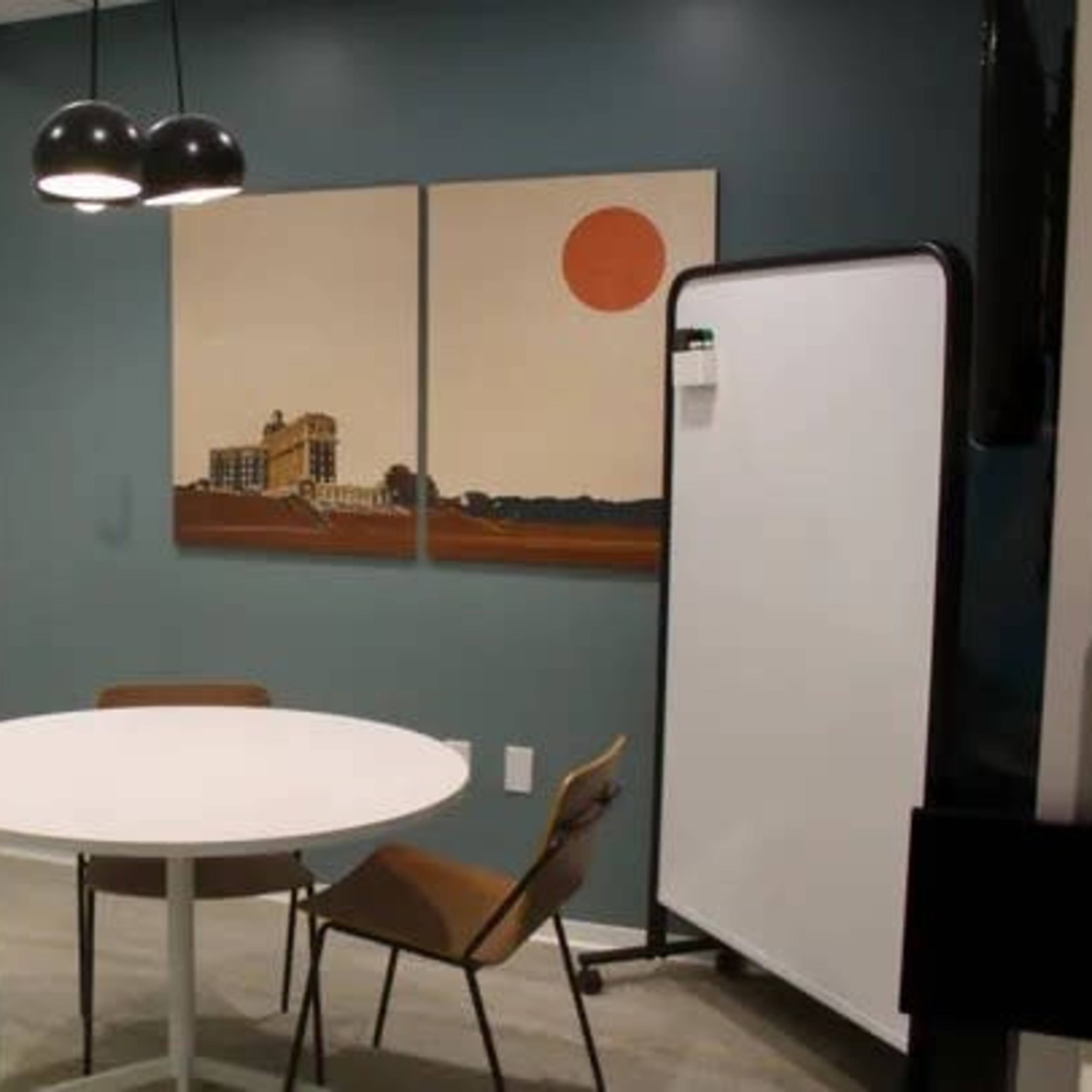 A modern office meeting room with a round table, two chairs, and a large glass wall featuring artwork and a whiteboard.