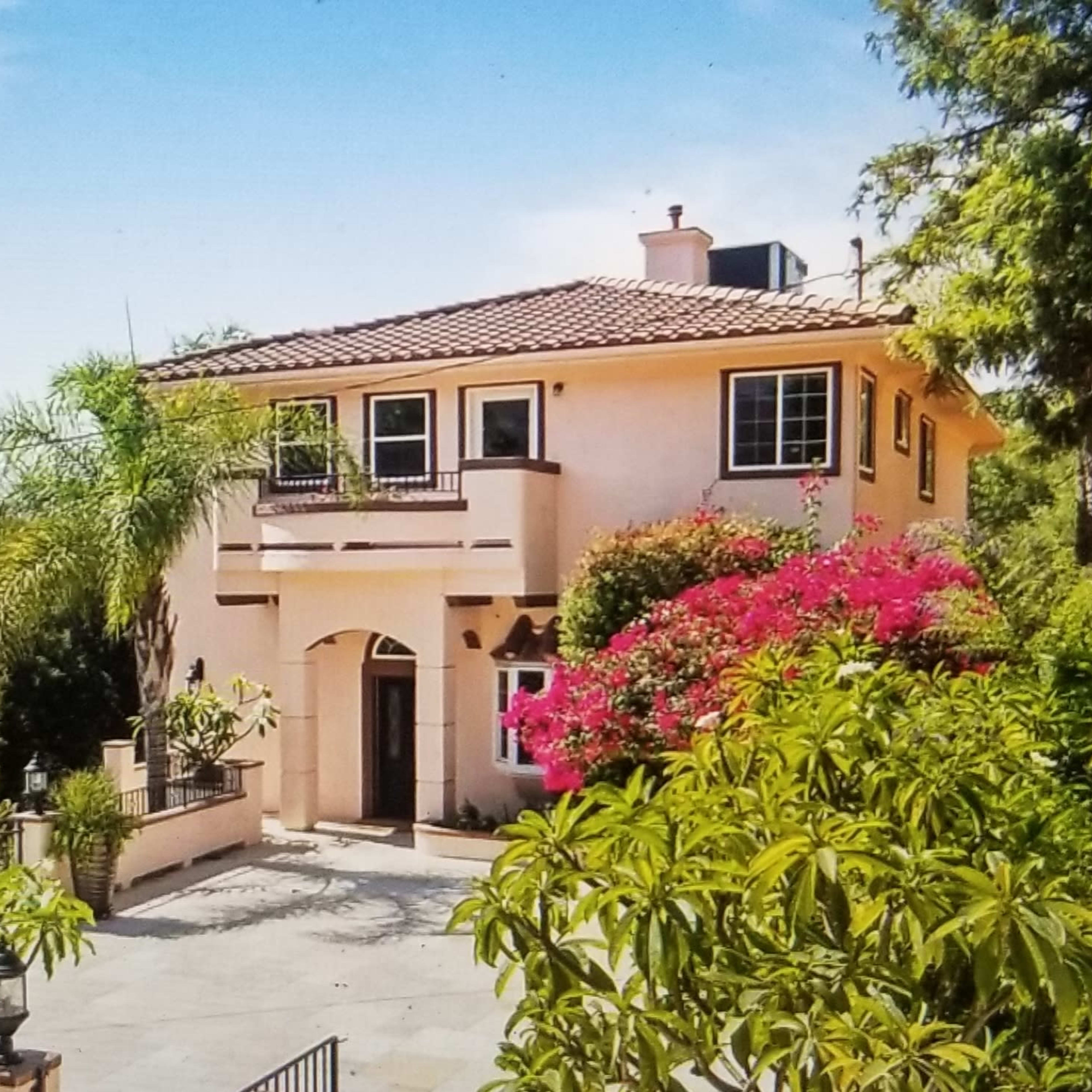 A two-story house with a terracotta roof is surrounded by lush greenery and colorful flowers.