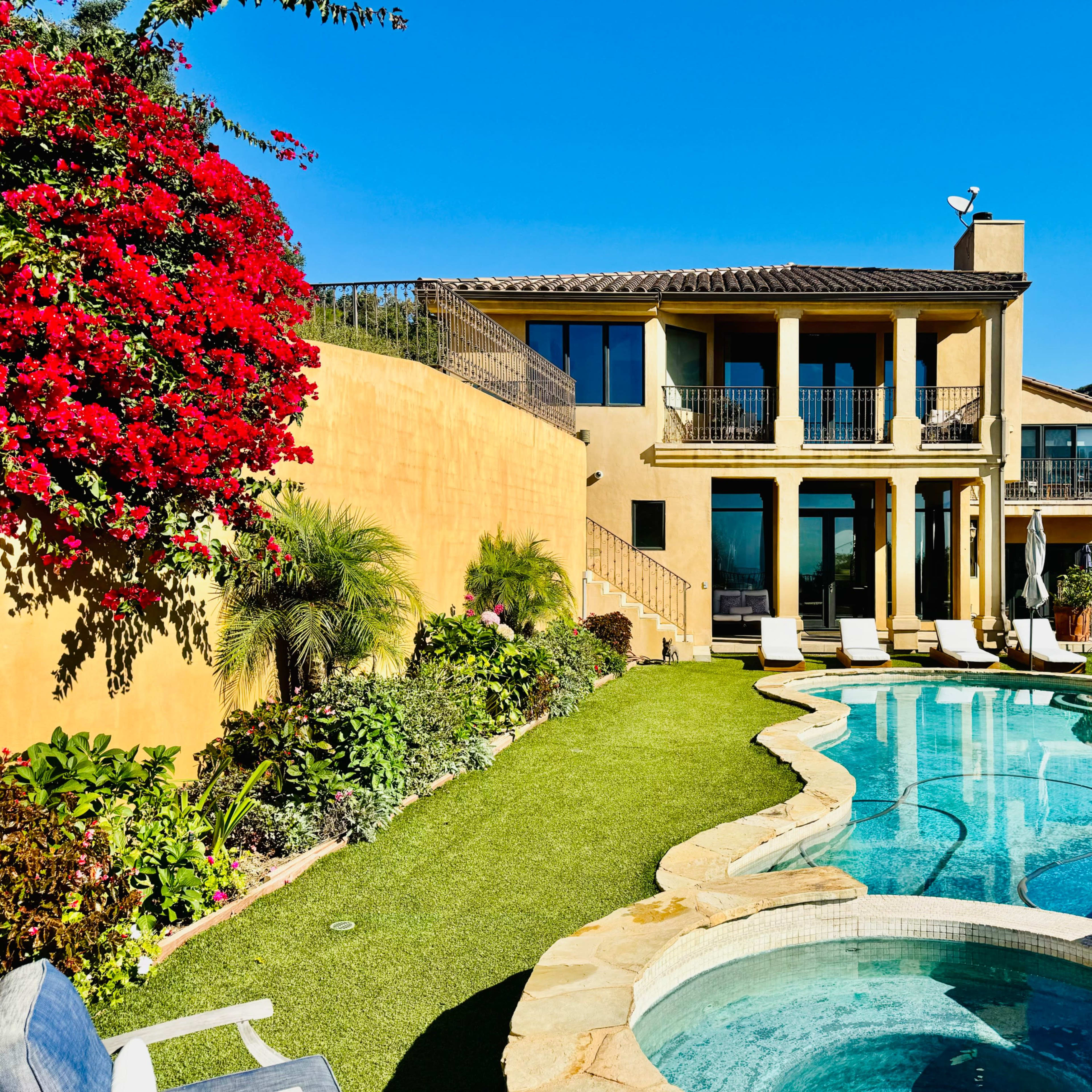 A well-maintained villa with a swimming pool and landscaped gardens, set against a clear blue sky.