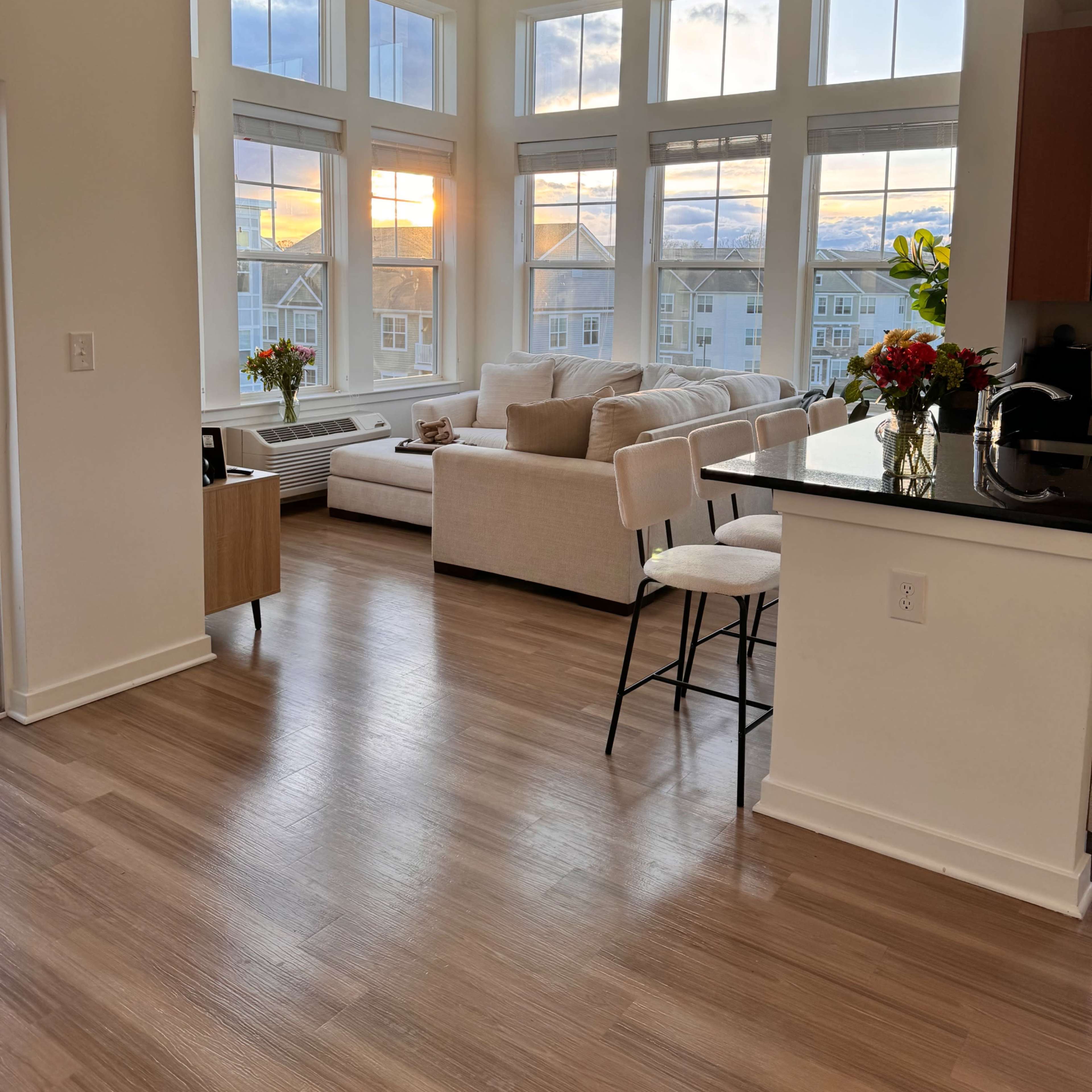 The scene depicts a modern living room with large windows overlooking a sunset, featuring a light-colored sectional sofa and a kitchen bar with stools.