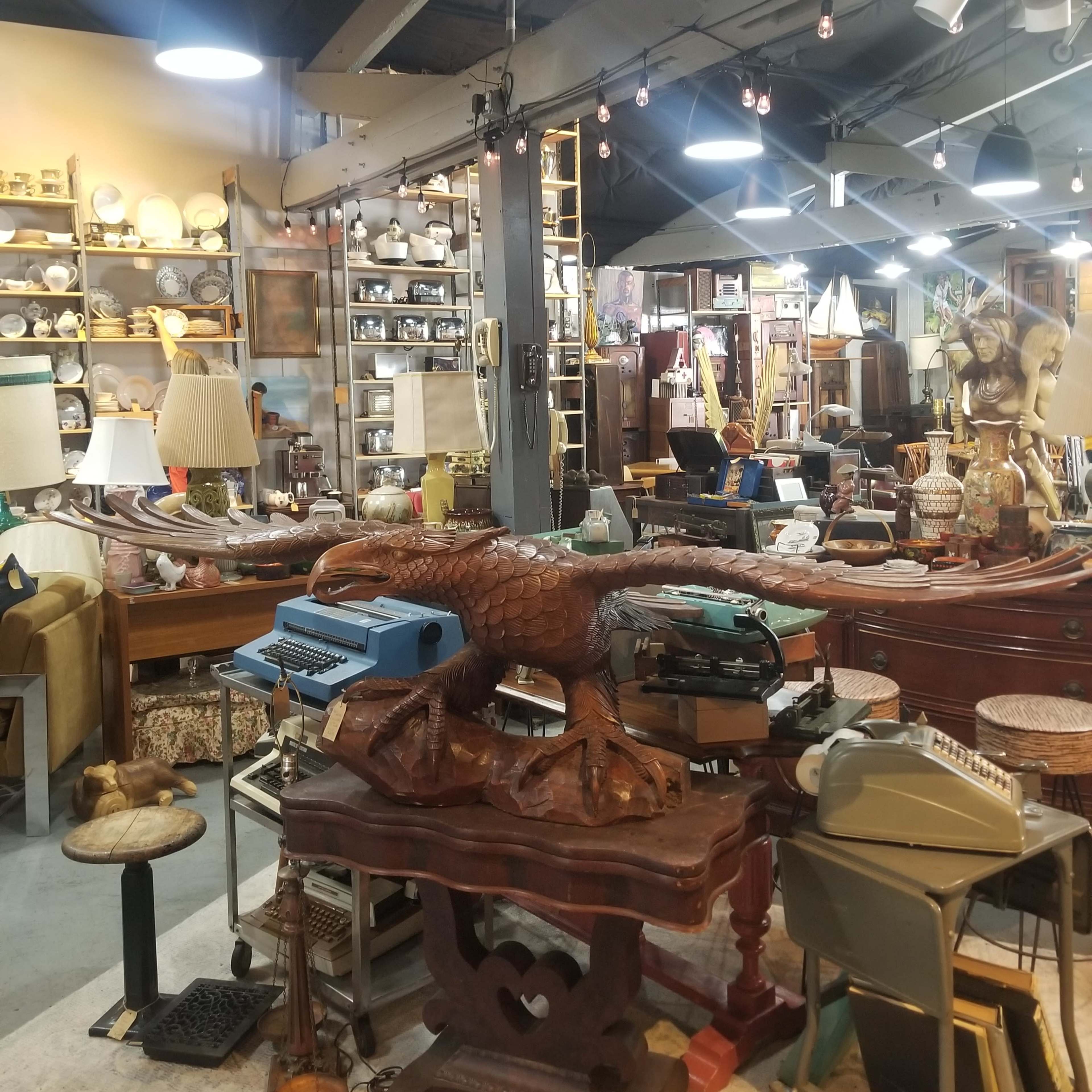 A cluttered antiques shop filled with a variety of vintage items, including furniture, lamps, and a typewriter, arranged across multiple displays.