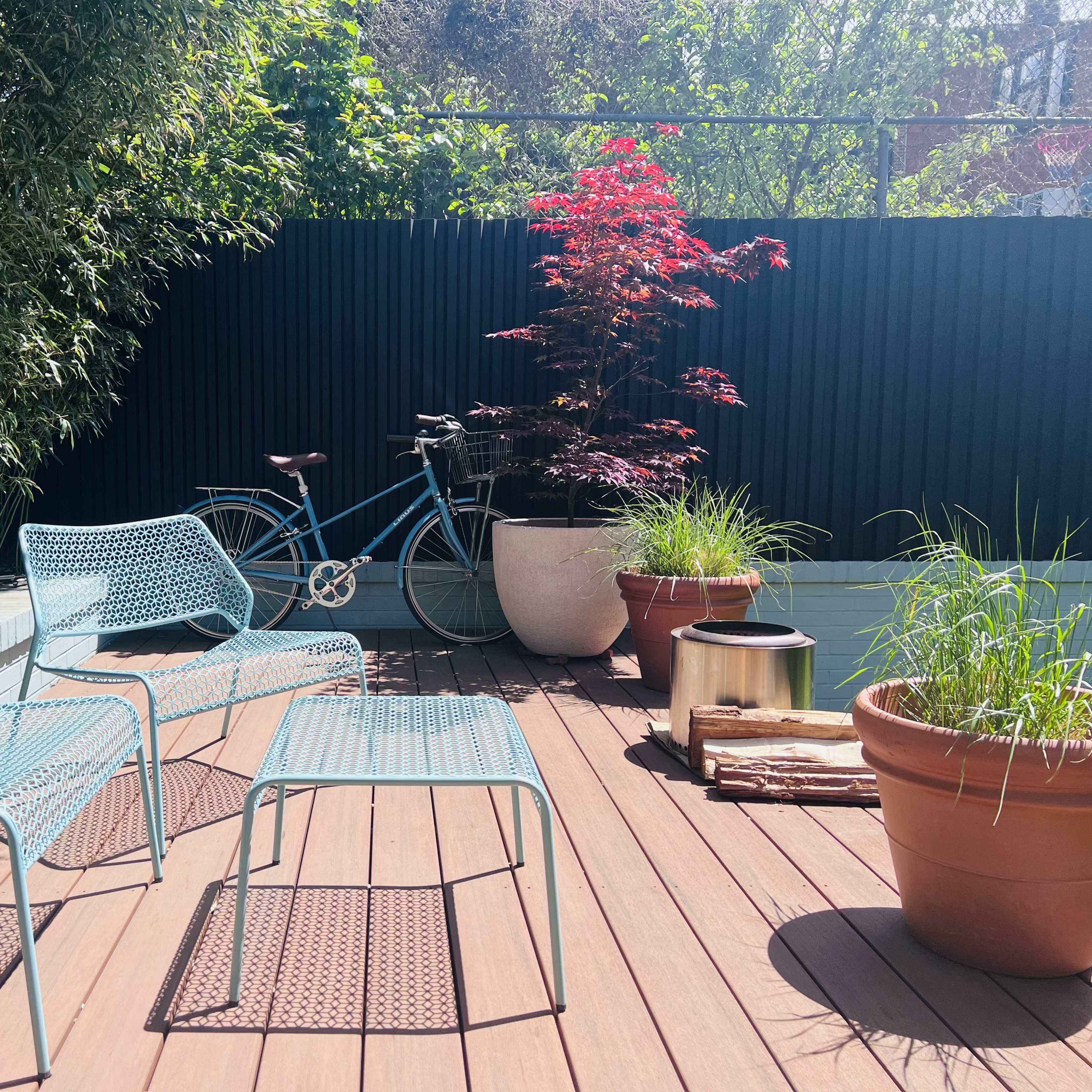 A small outdoor patio features metal chairs, a bicycle leaning against a black fence, and potted plants arranged on a wooden deck.