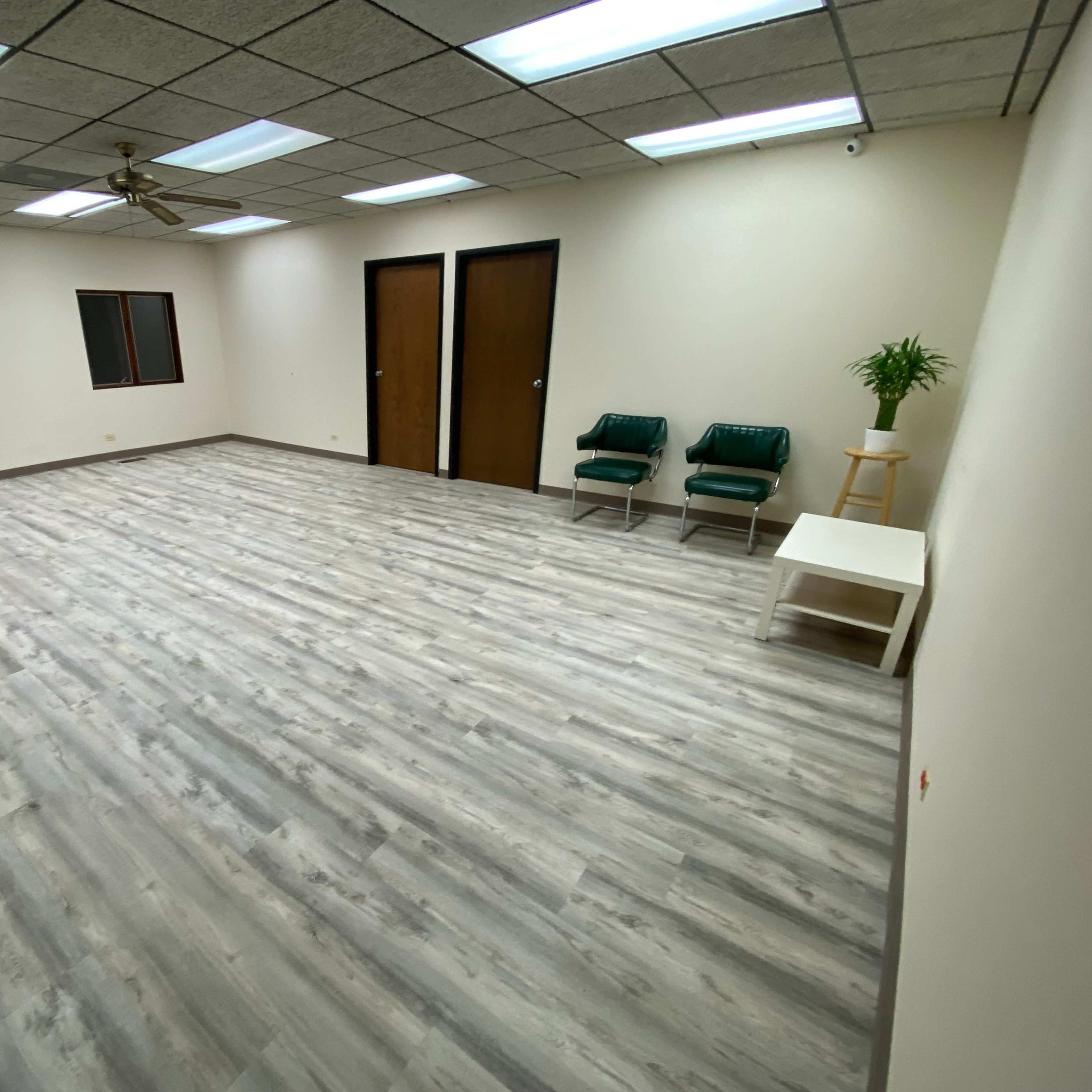 A spacious, empty room with light-colored wooden flooring, two green chairs, a small white table, and a mirror against the wall.