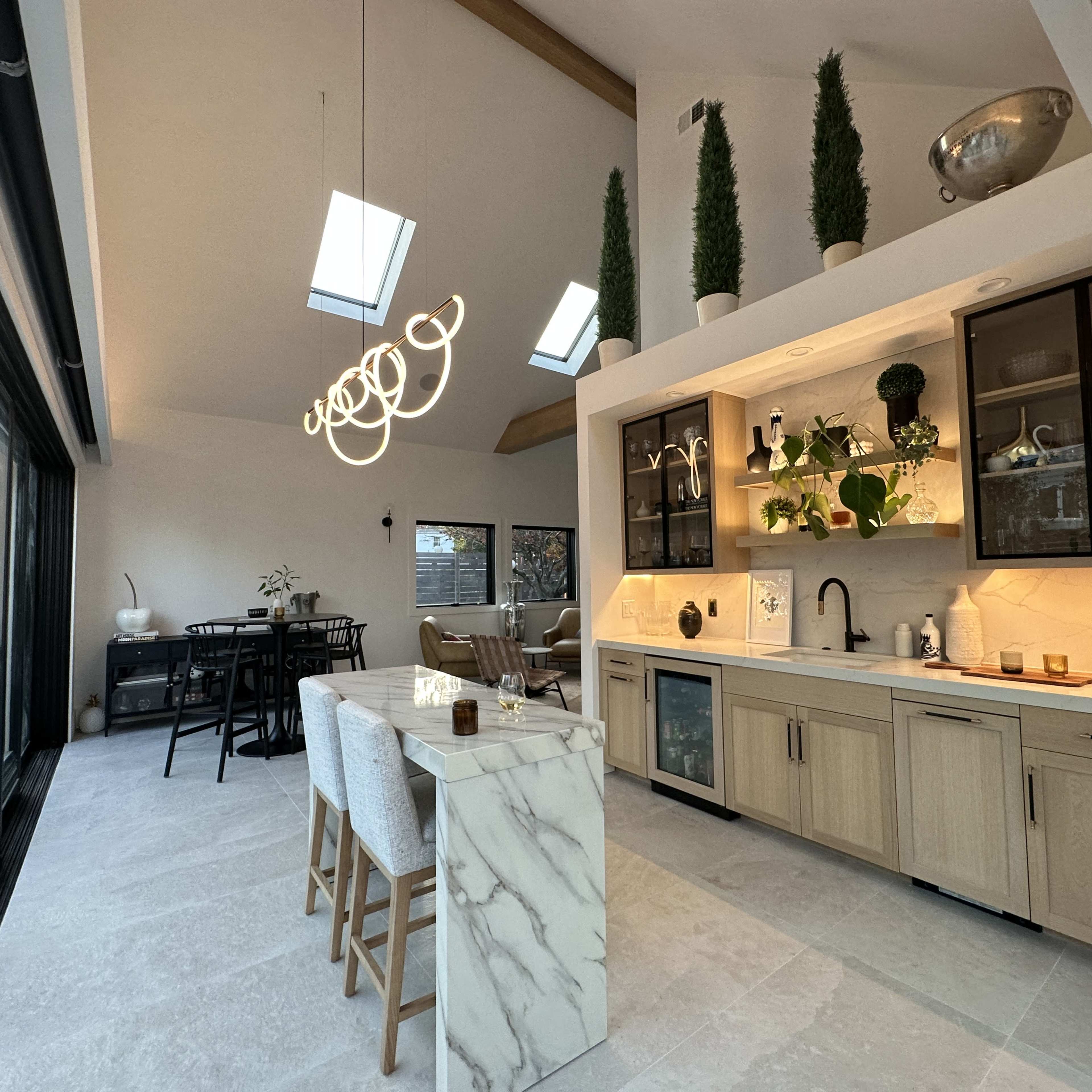 The scene shows a modern kitchen and dining area featuring a central marble island, wooden cabinetry, and large windows with a skylight.