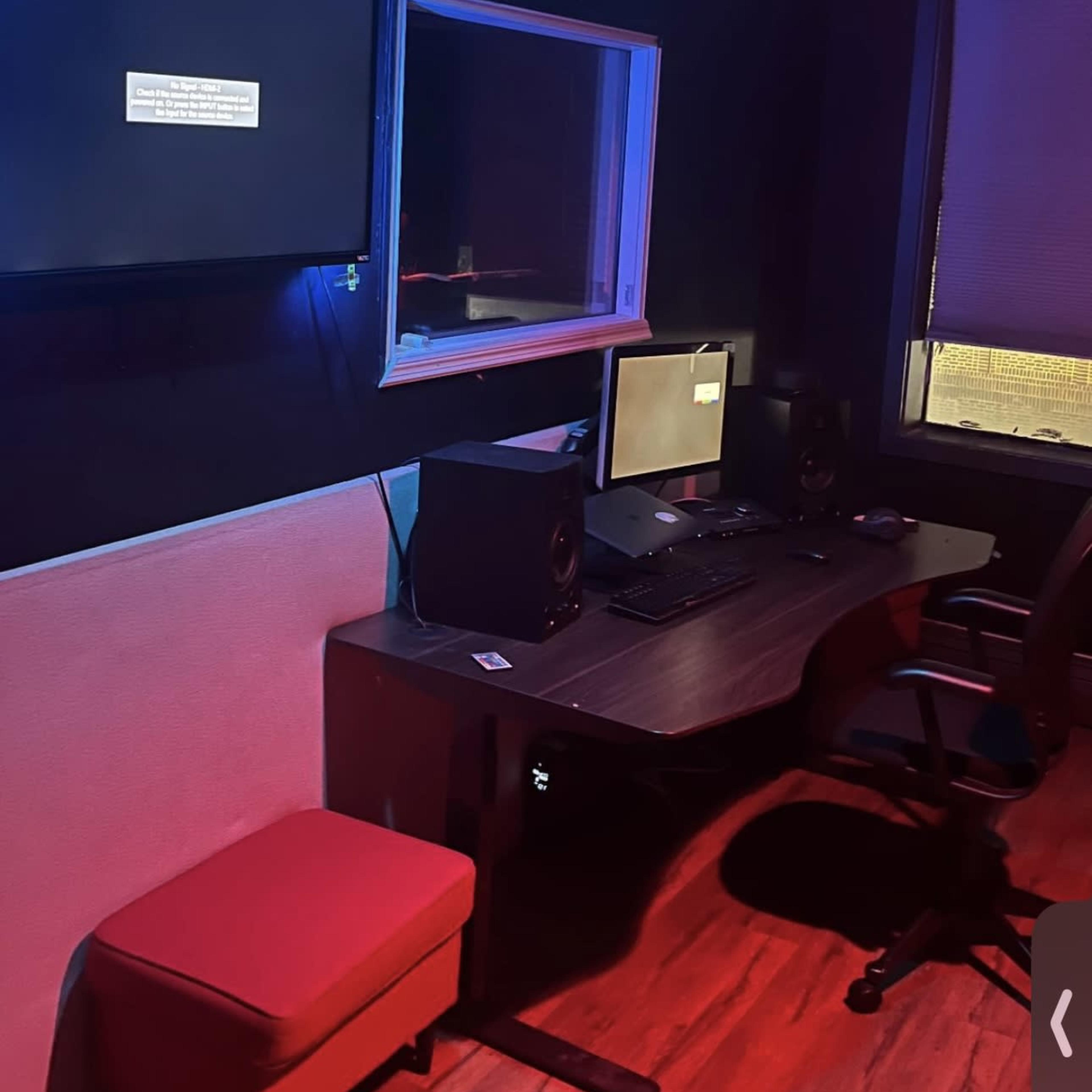 A dimly lit room contains a desk with two computer monitors, speakers, and a red chair, along with a wall-mounted screen displaying a message.