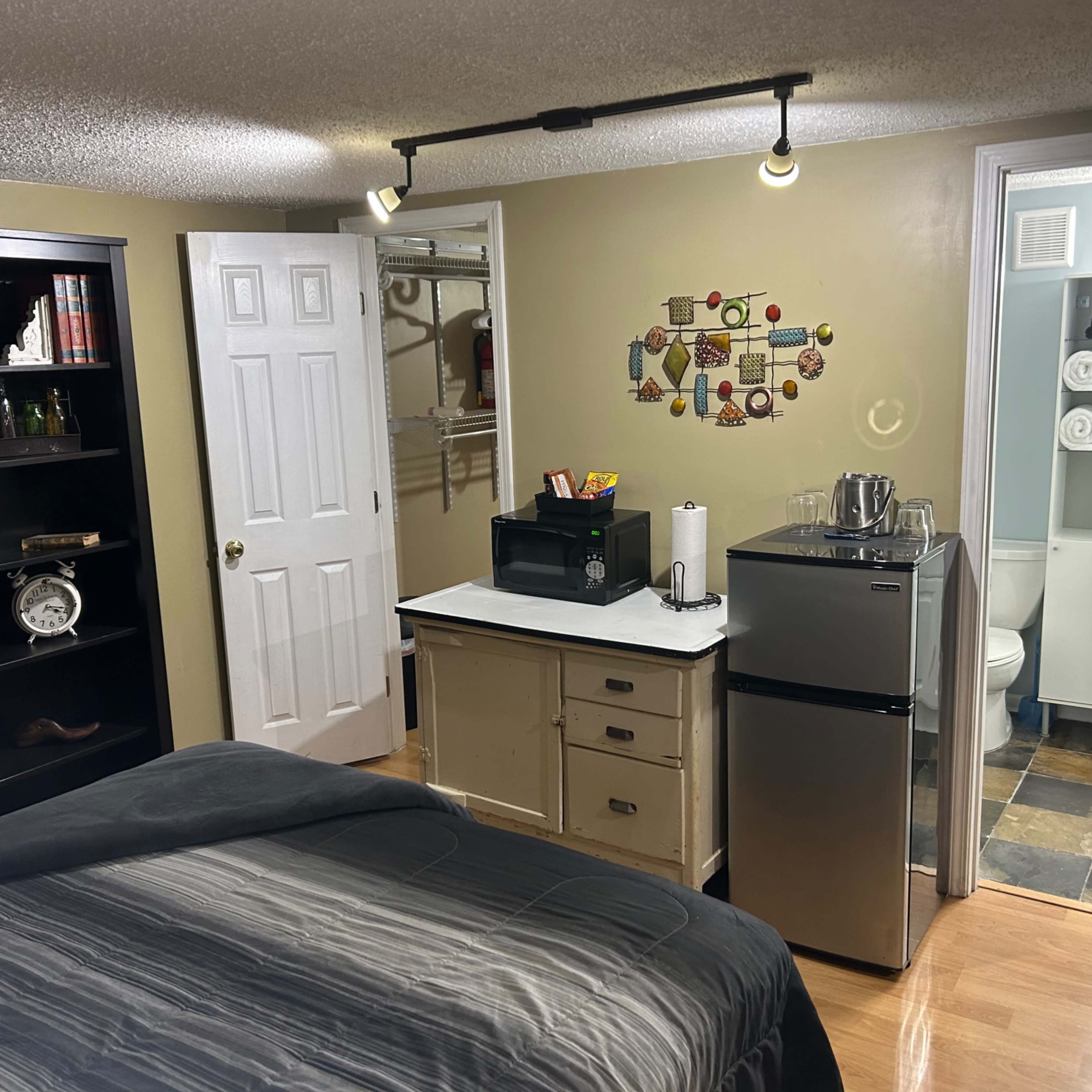 A small bedroom with a black bookshelf, a queen-sized bed, a mini fridge, and a workspace area, along with a bathroom visible through an open door.
