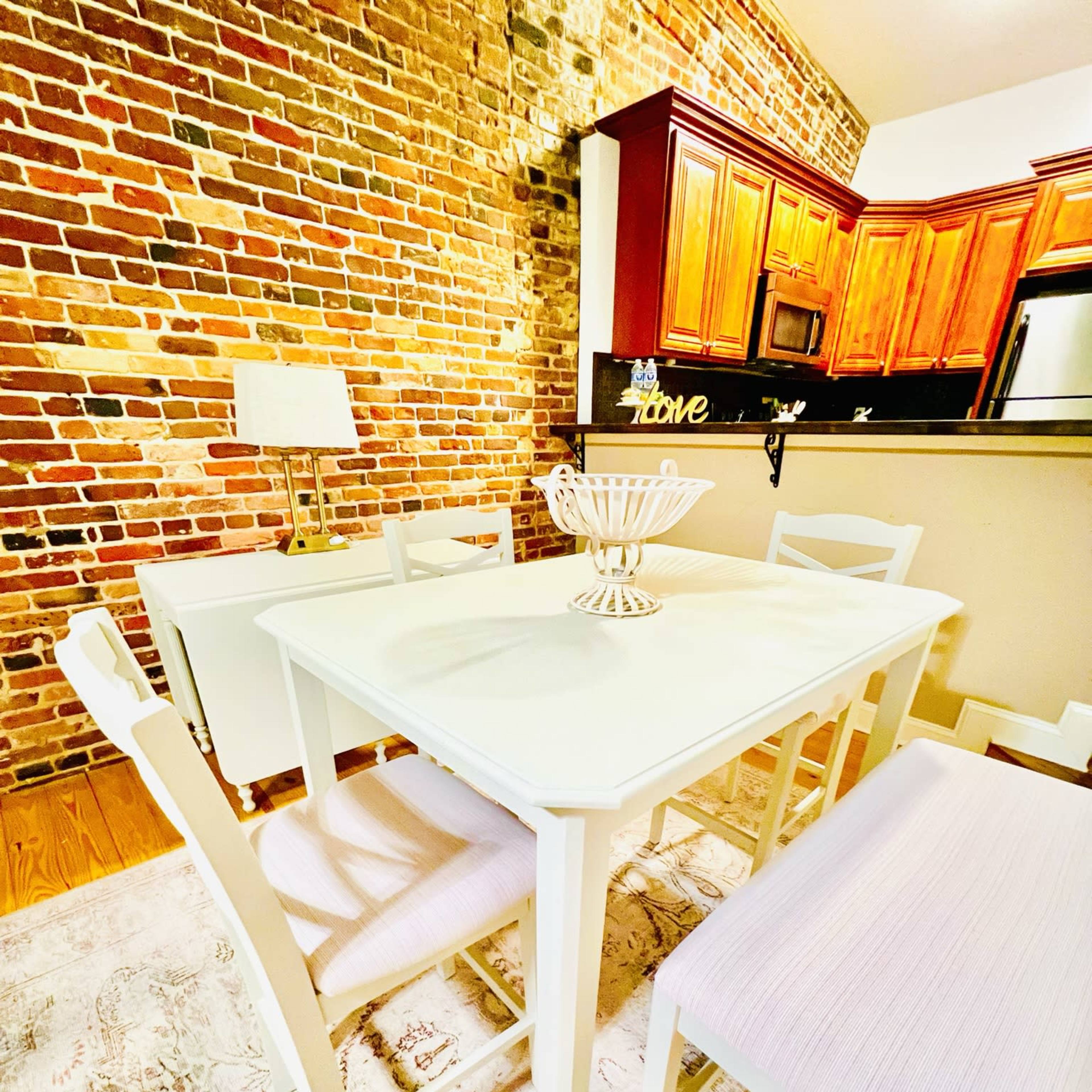 A white dining table surrounded by wooden chairs on a patterned rug, with a brick wall and kitchen cabinetry in the background.