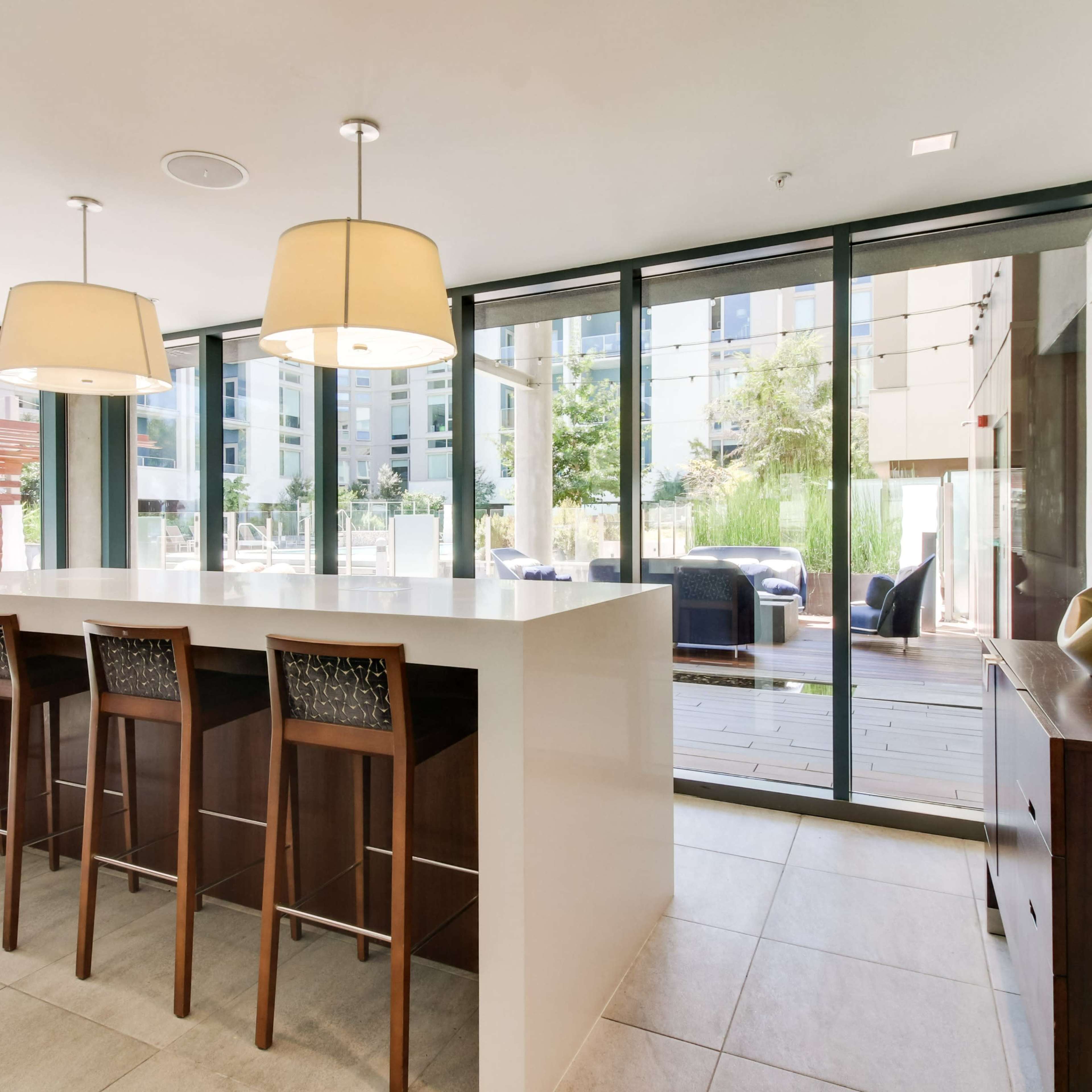 A modern kitchen features a large white island with three wooden bar stools and large windows overlooking an outdoor patio area.
