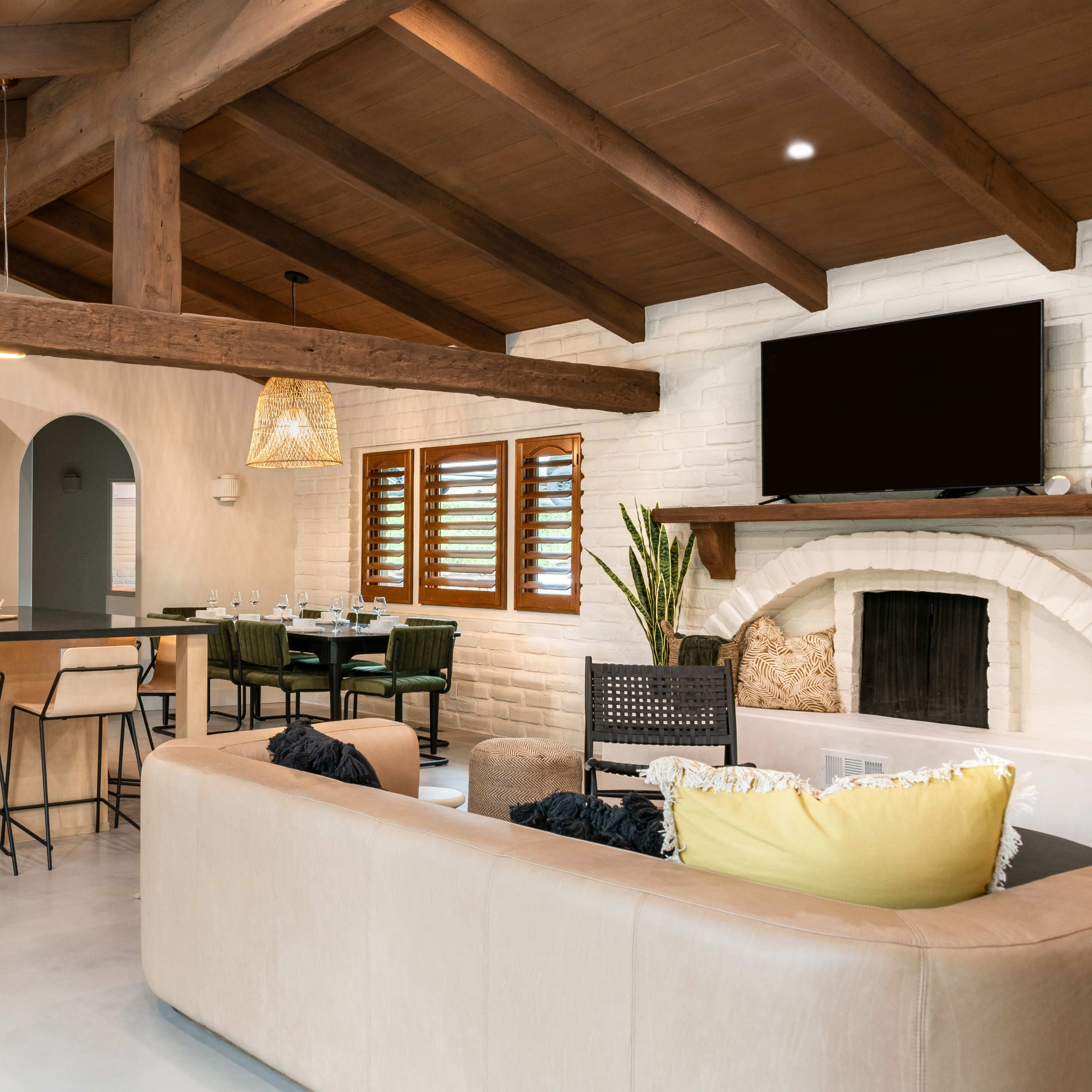 A modern kitchen and living area featuring wooden beams, a large island with bar stools, and a cozy seating arrangement with a television mounted on the wall.