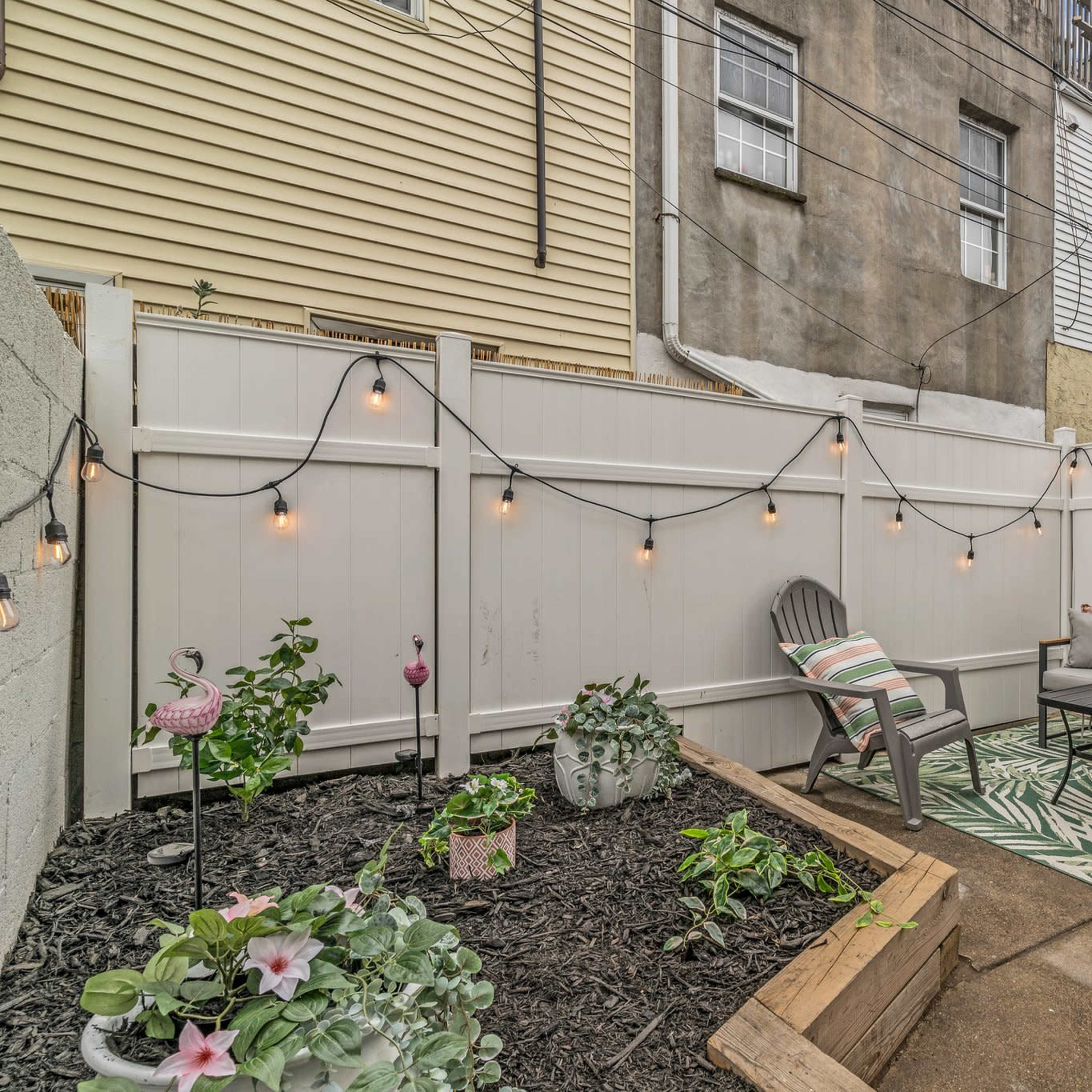 A small outdoor patio area with a couch, coffee table, and string lights, surrounded by a white fence and a garden bed with plants.