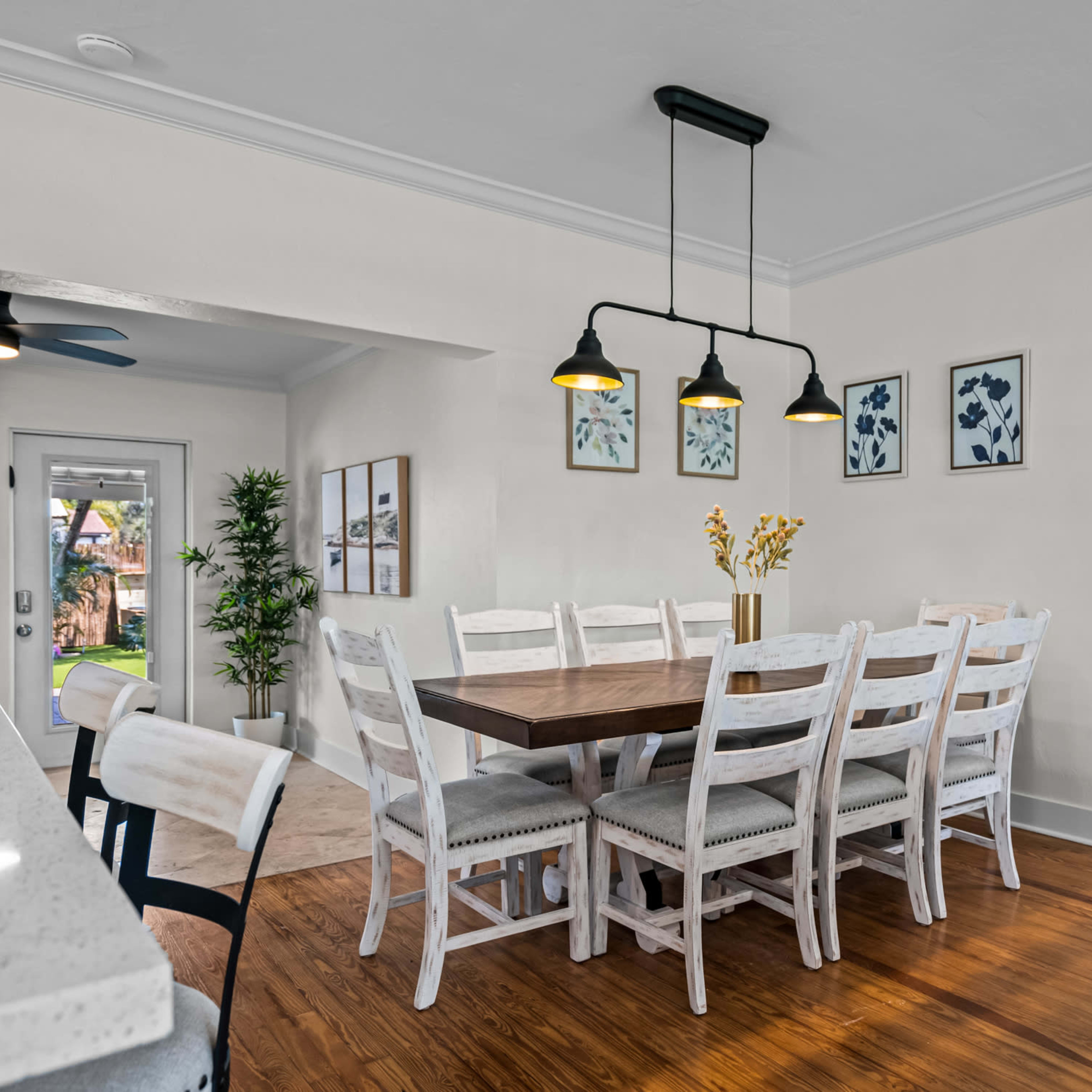 A dining area features a large wooden table surrounded by white chairs, illuminated by pendant lights, with framed artwork on the walls.