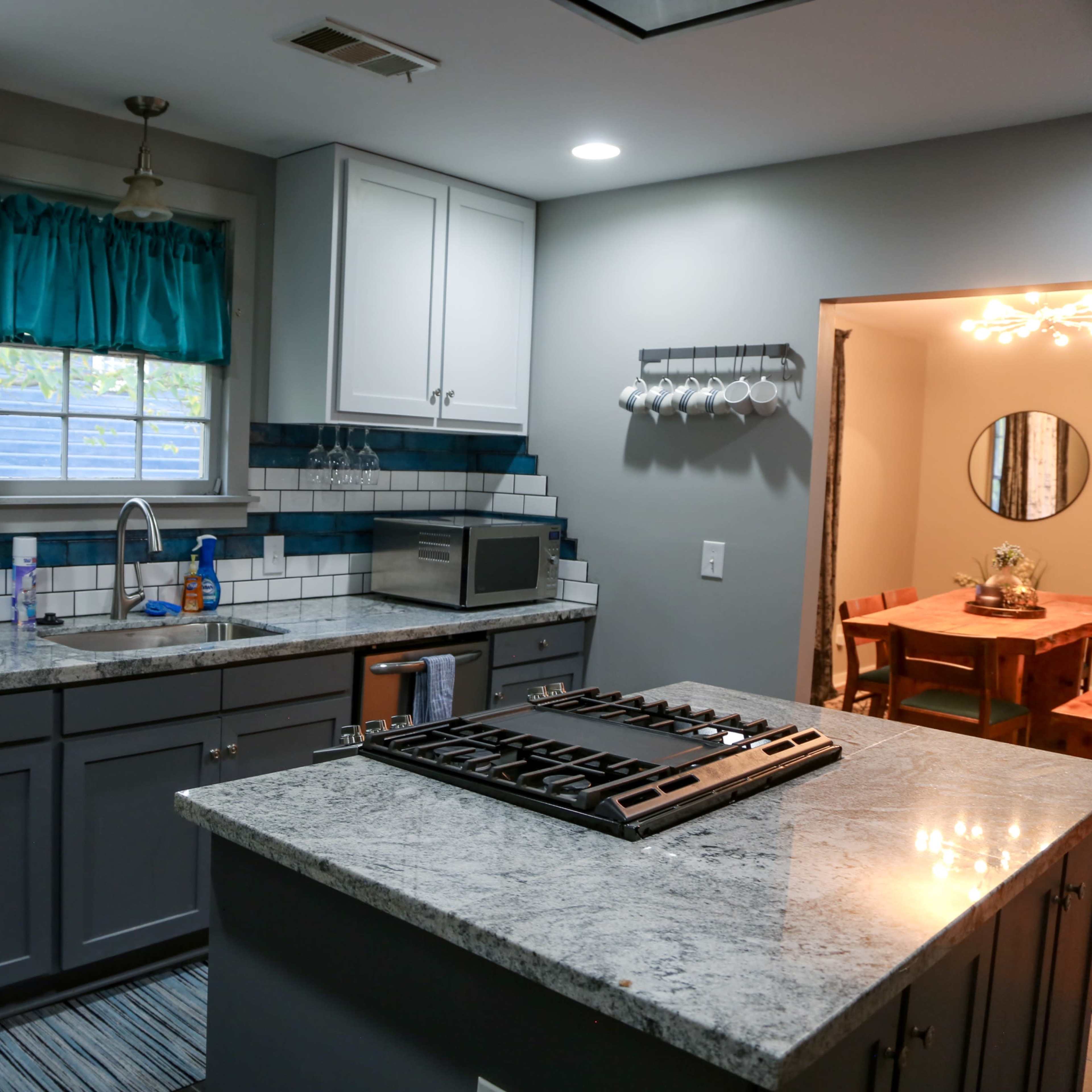 A modern kitchen features gray cabinets, a granite countertop, and stainless steel appliances, with a dining area visible through an adjacent doorway.