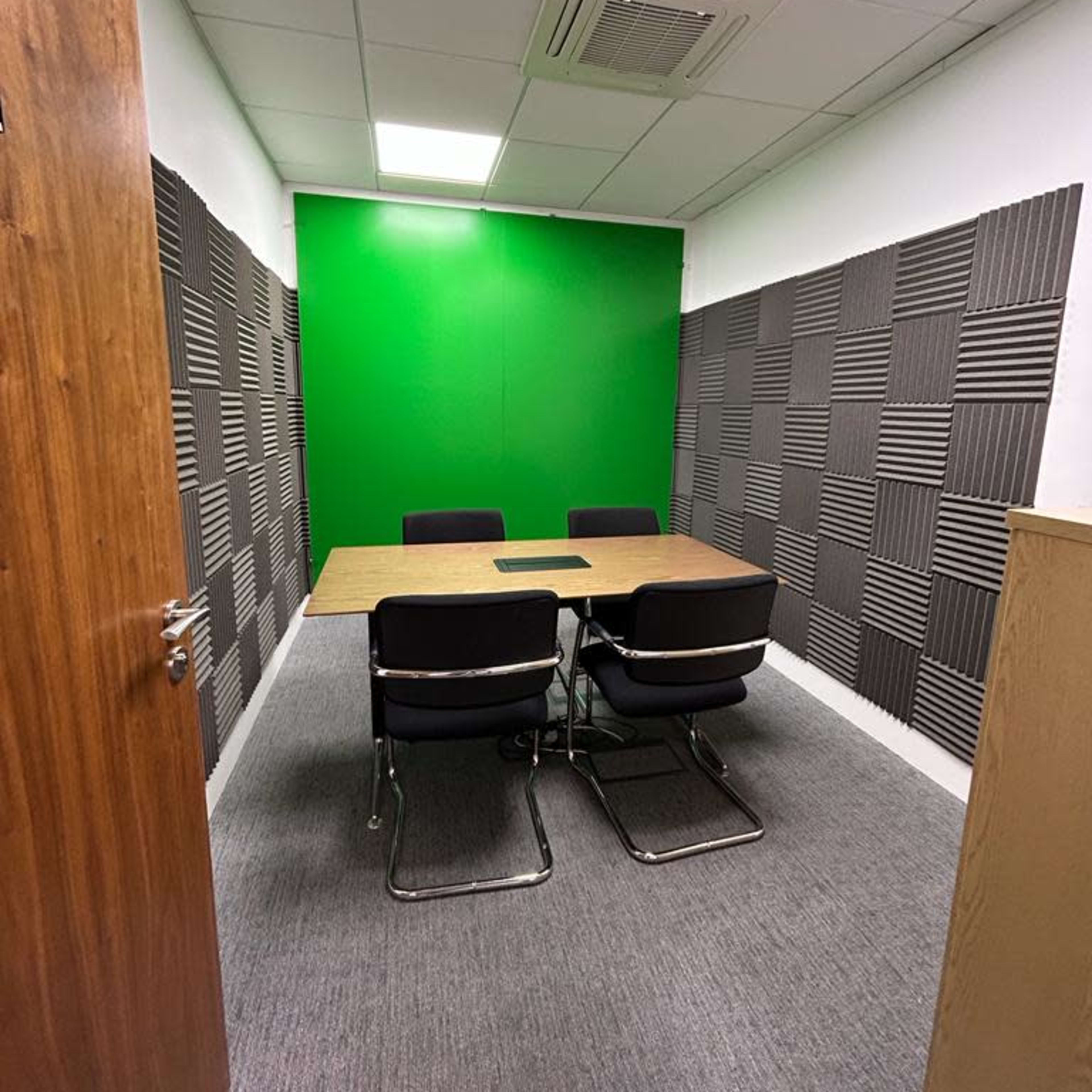 A small meeting room with a wooden table and four black chairs, featuring soundproof panels on the walls and a bright green backdrop.
