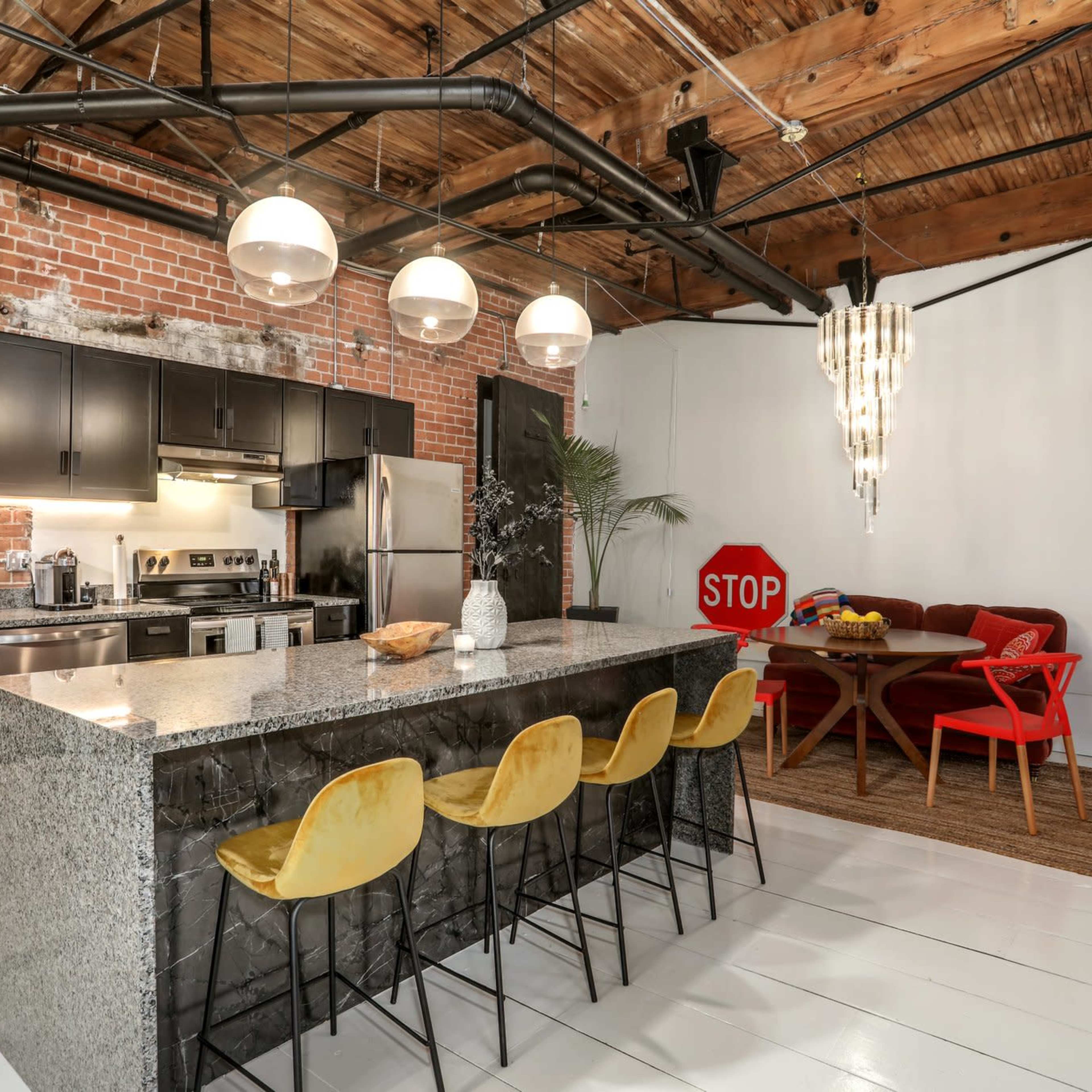 A modern kitchen with a granite island and bar seating, featuring exposed brick walls and industrial-style lighting, alongside a dining area with a red table and chairs.
