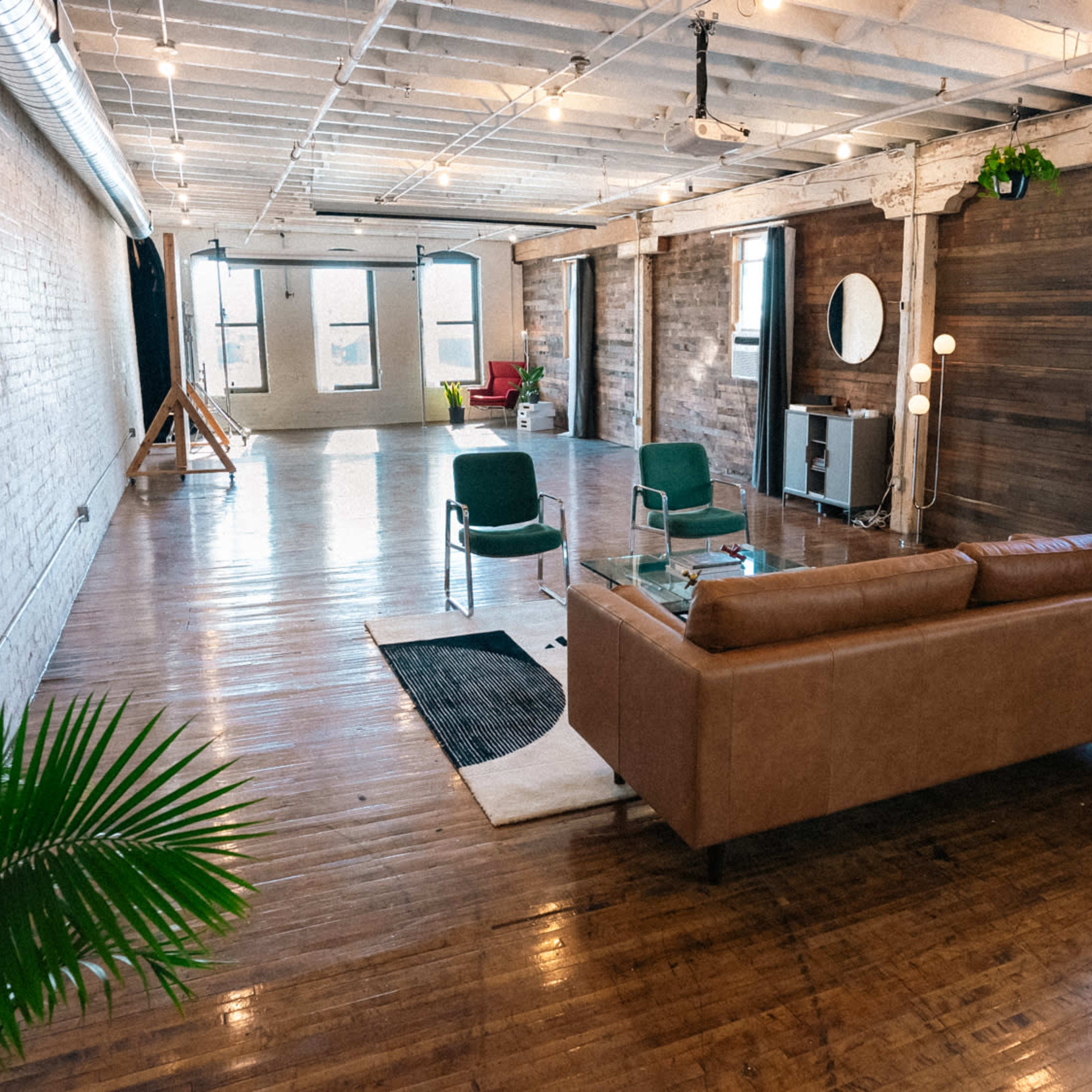 A spacious interior with hardwood floors, featuring a brown sofa, green chairs, a plant, and large windows that illuminate the room.