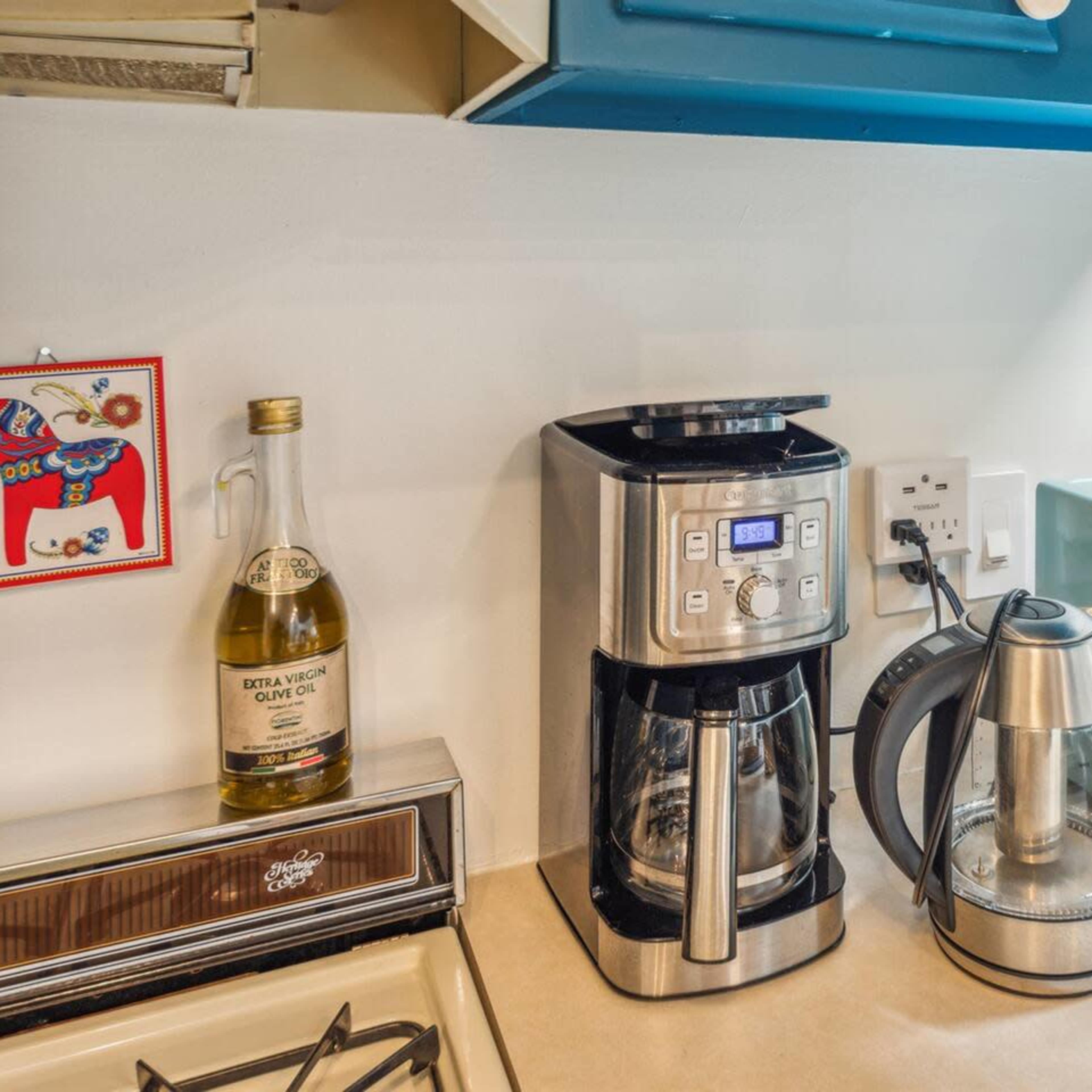 A kitchen countertop with a coffee maker, an electric kettle, a blue toaster oven, a bottle of olive oil, and a gas stove.