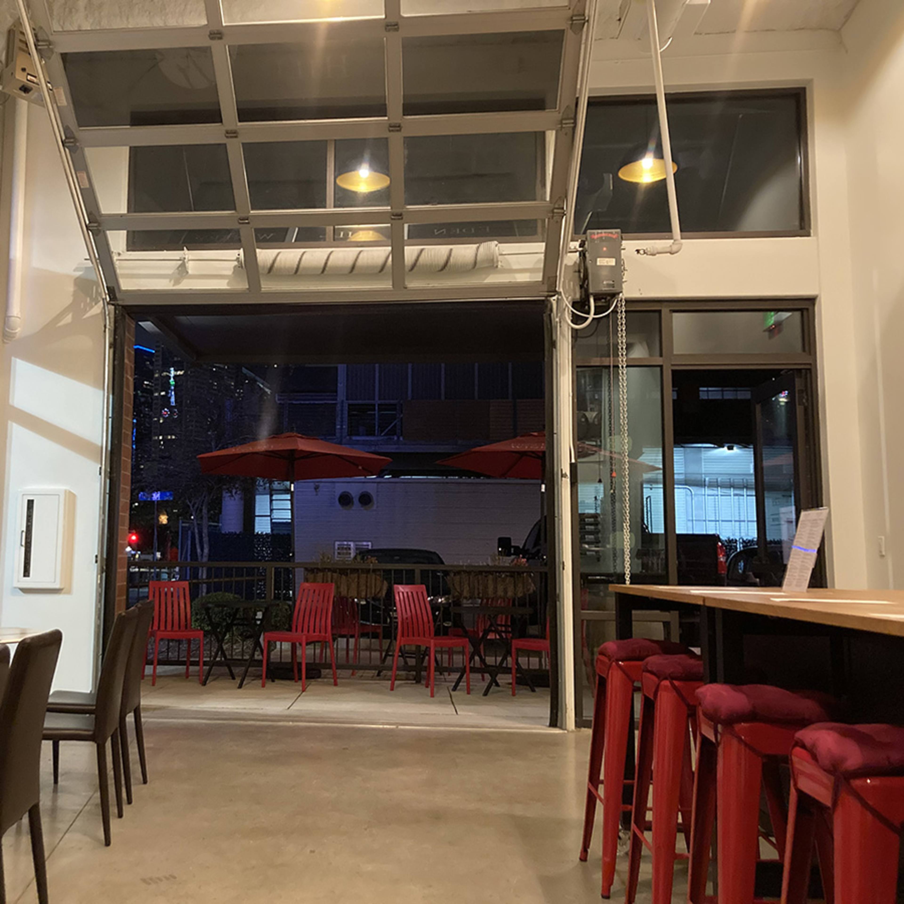 An interior space with a large open garage door that leads to an outdoor seating area, featuring red chairs and umbrellas.
