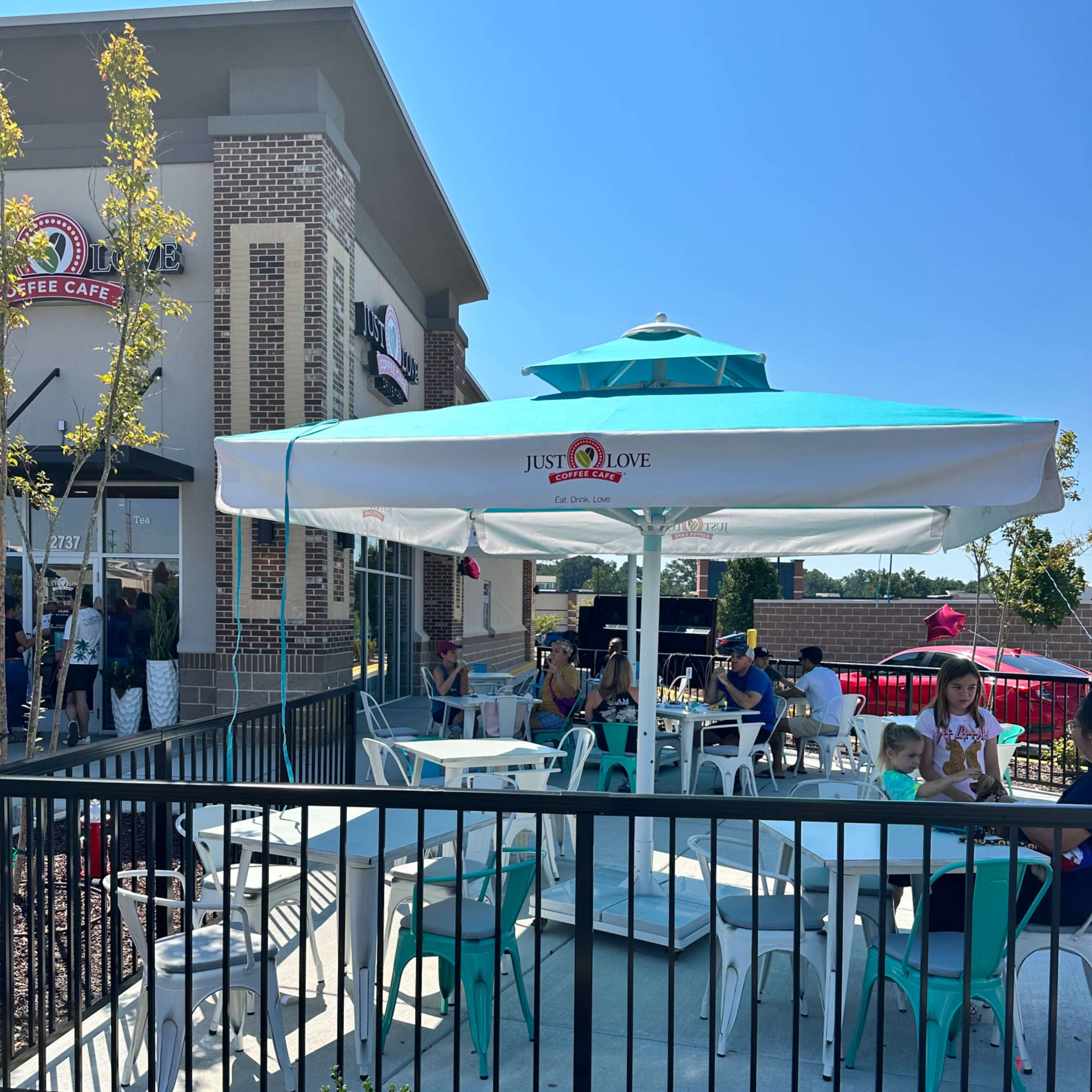 A patio area with white tables and turquoise chairs is situated outside a café, shaded by a large umbrella.