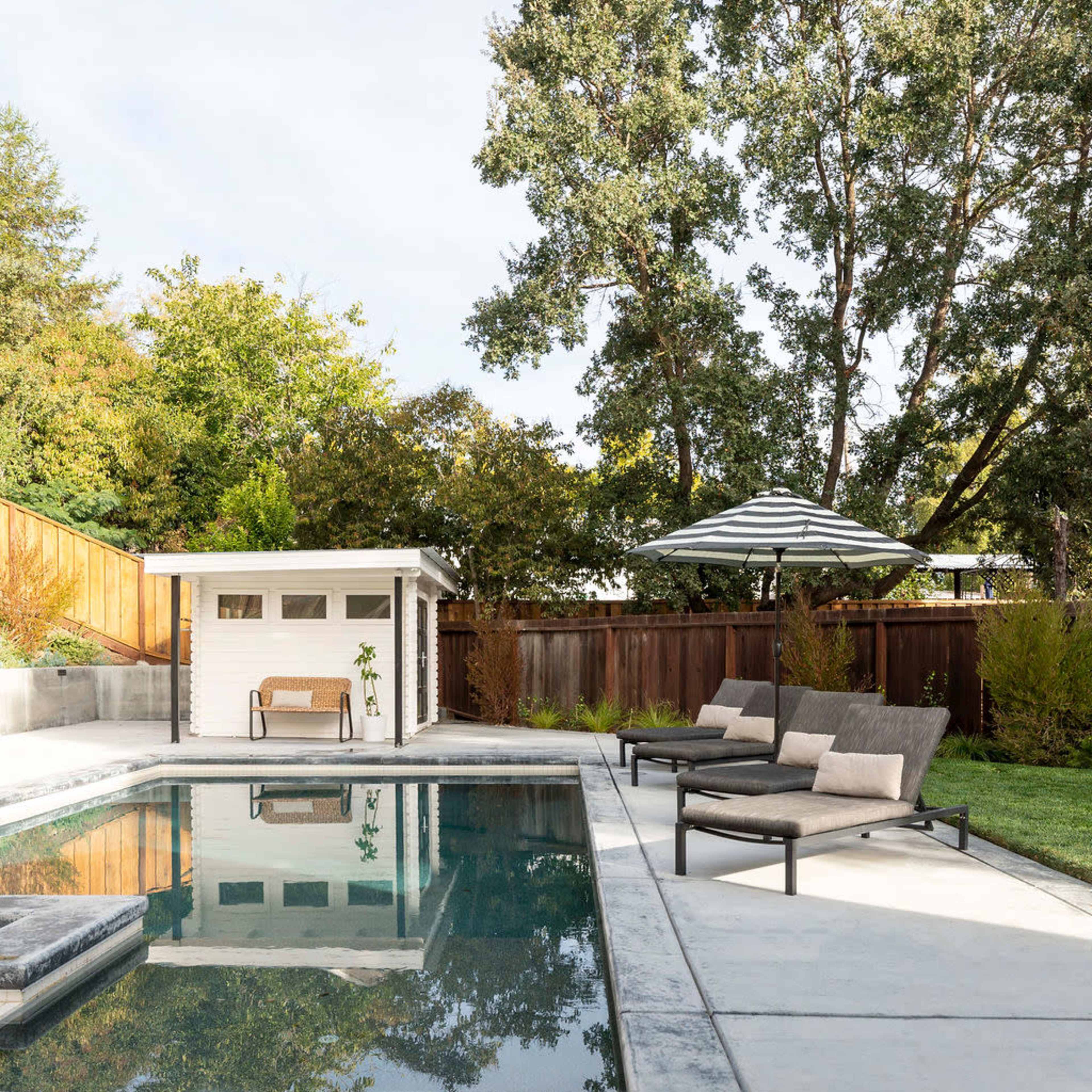 A serene backyard features a swimming pool, lounge chairs, and a white shed surrounded by trees and greenery.