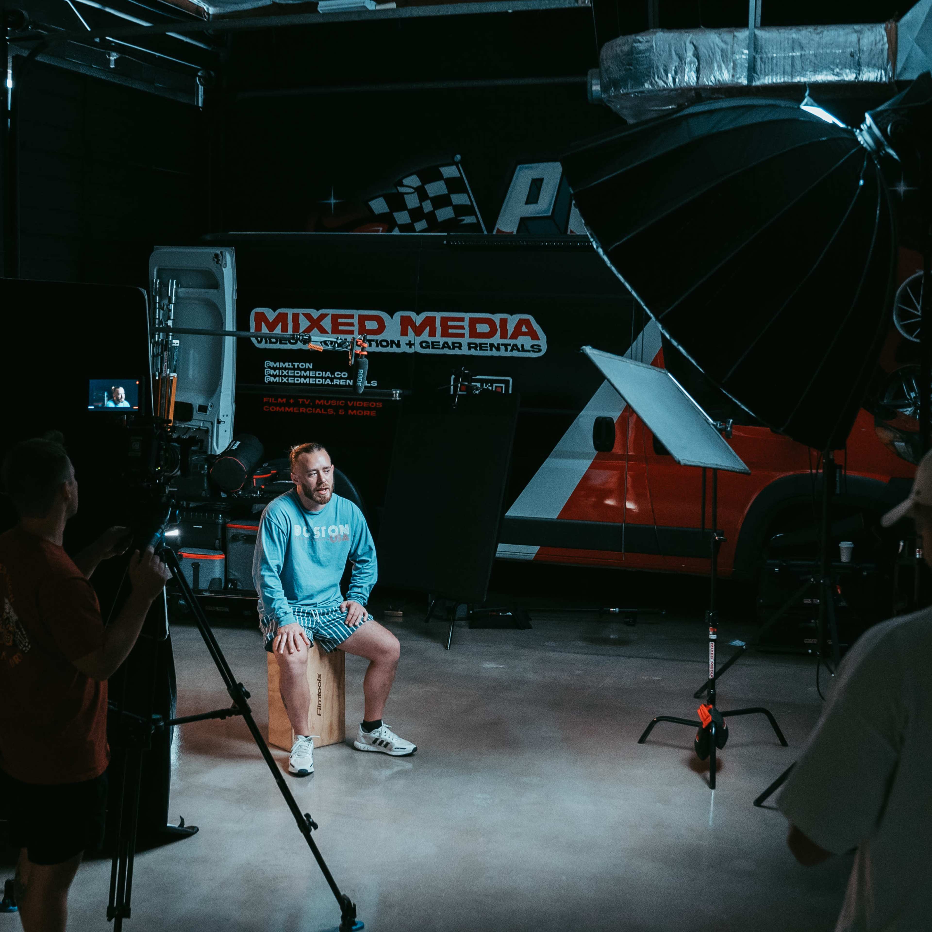 A man sits on a wooden stool in a studio, surrounded by camera equipment and lighting setups.