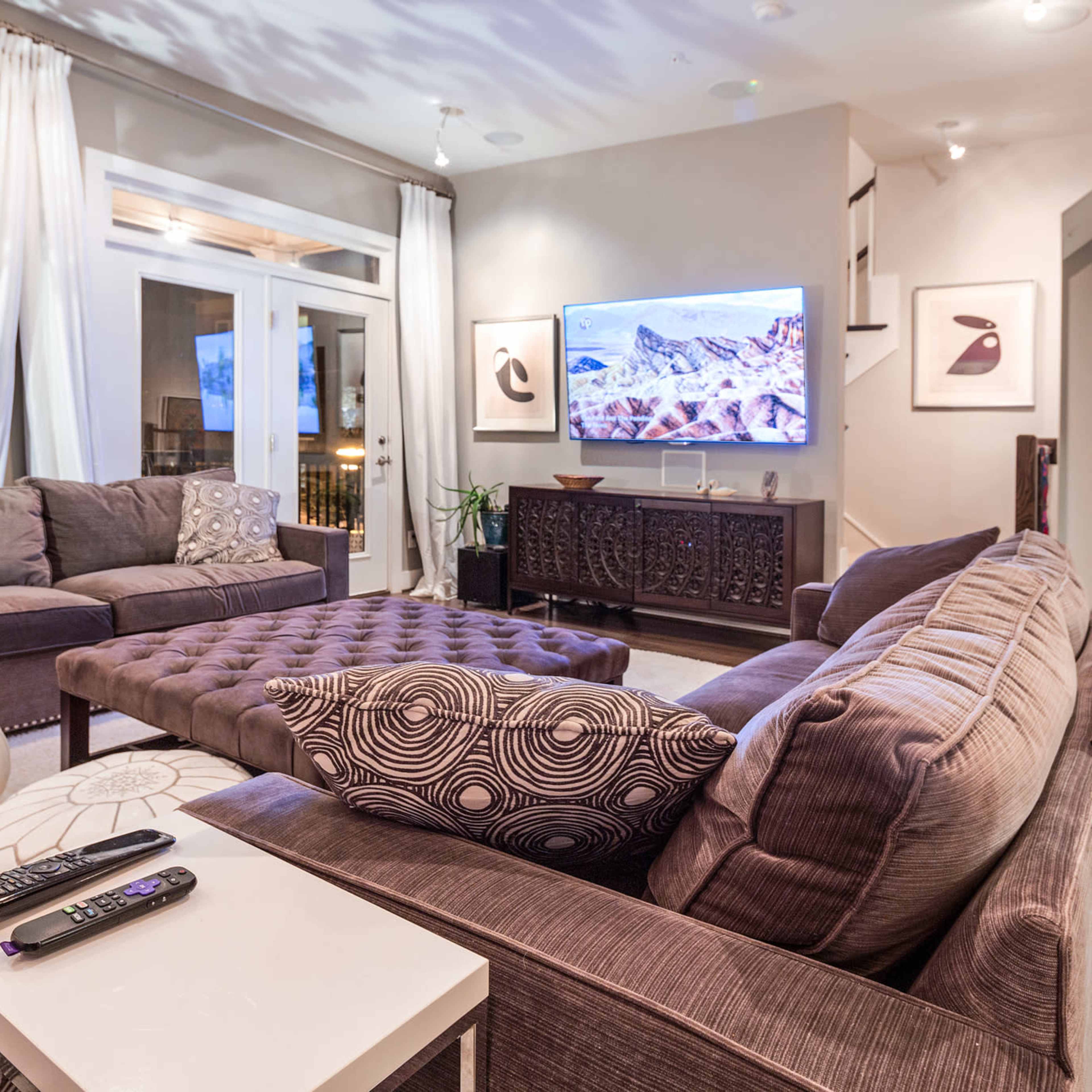 The living room features a large sectional sofa, an ottoman, and a wall-mounted television, with sheer curtains and decorative items enhancing the space.