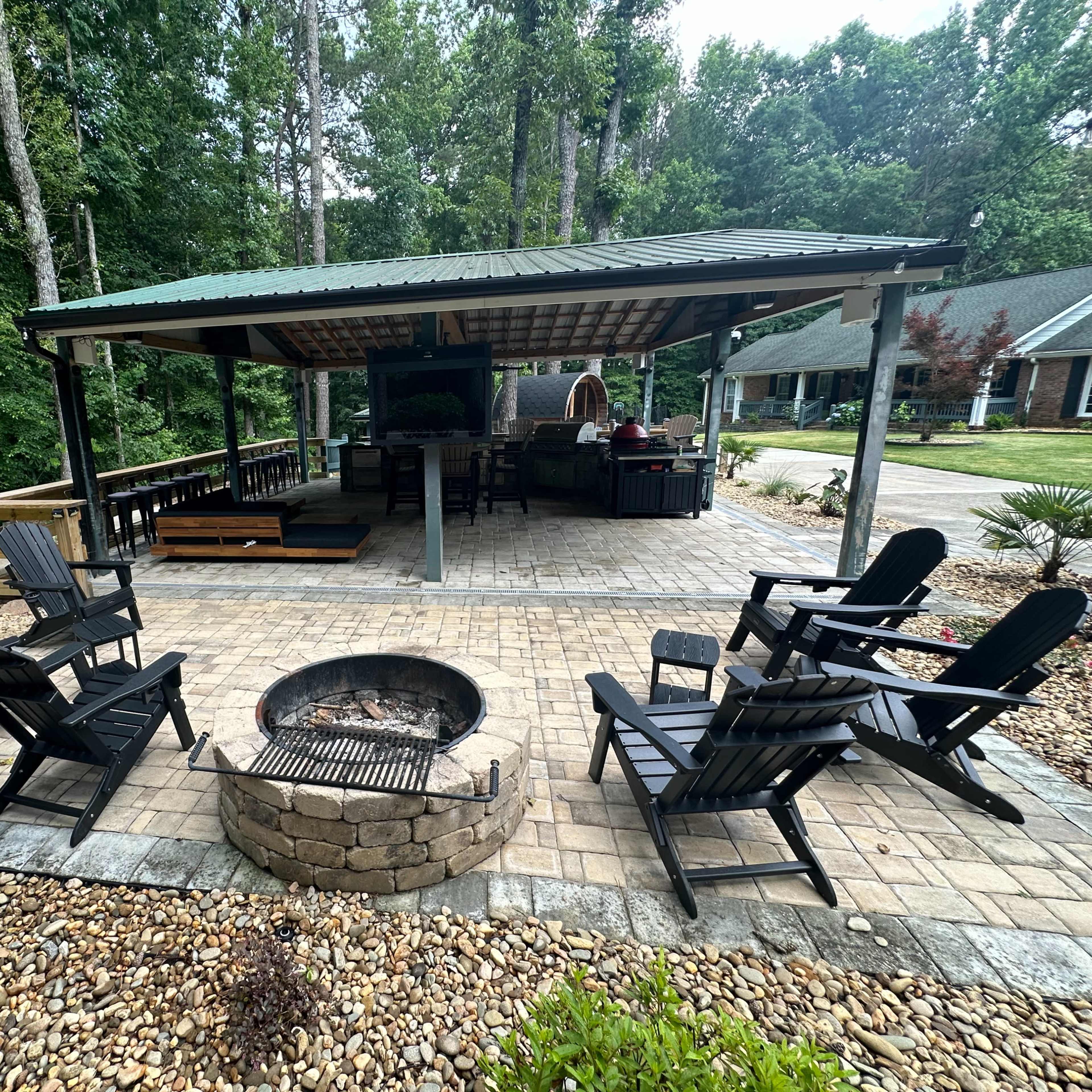 A covered outdoor area features a stone patio with a fire pit surrounded by black chairs and a grill, set in a wooded backdrop.