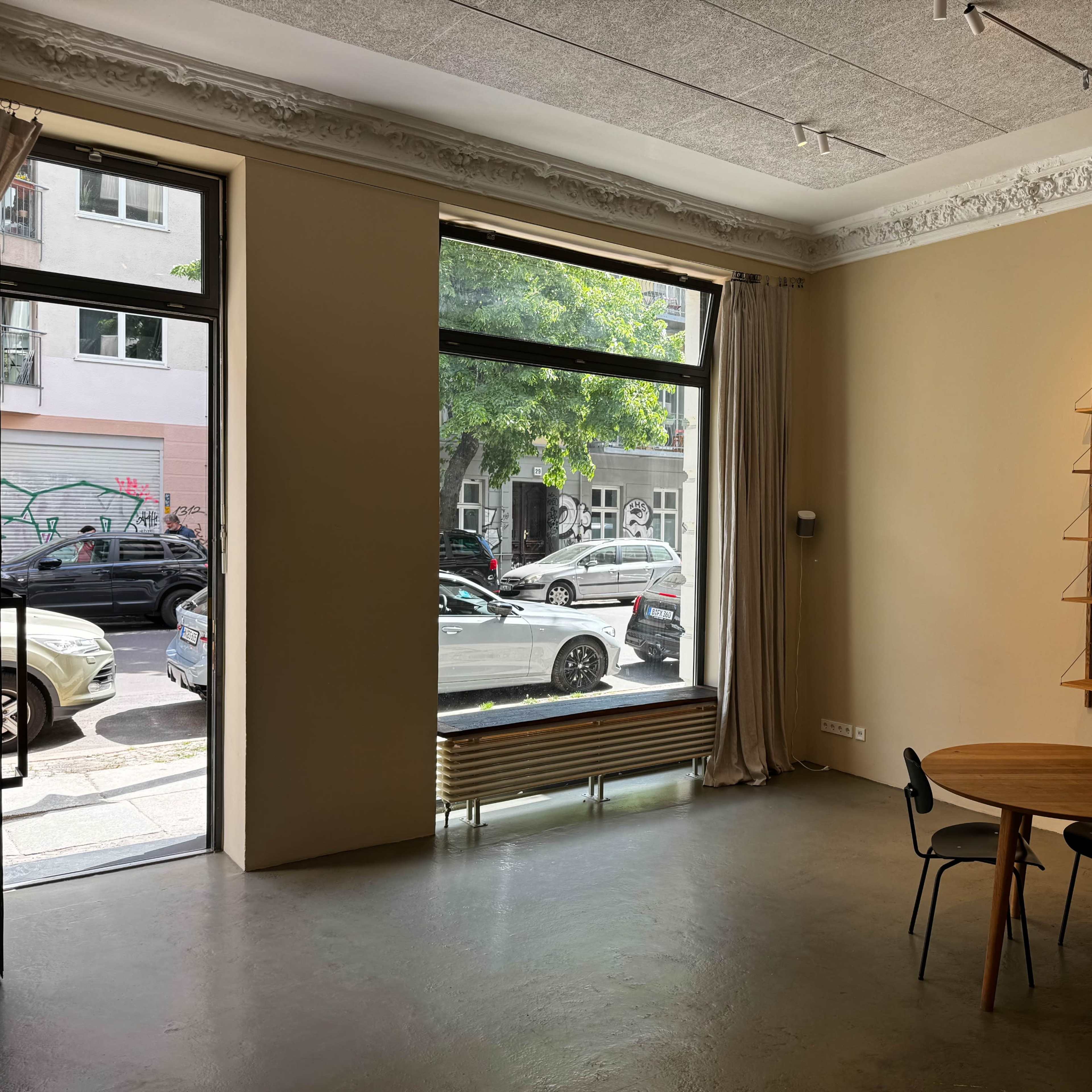 A minimalist interior space with a round wooden table and two chairs, featuring large windows that open to a view of parked cars and greenery outside.