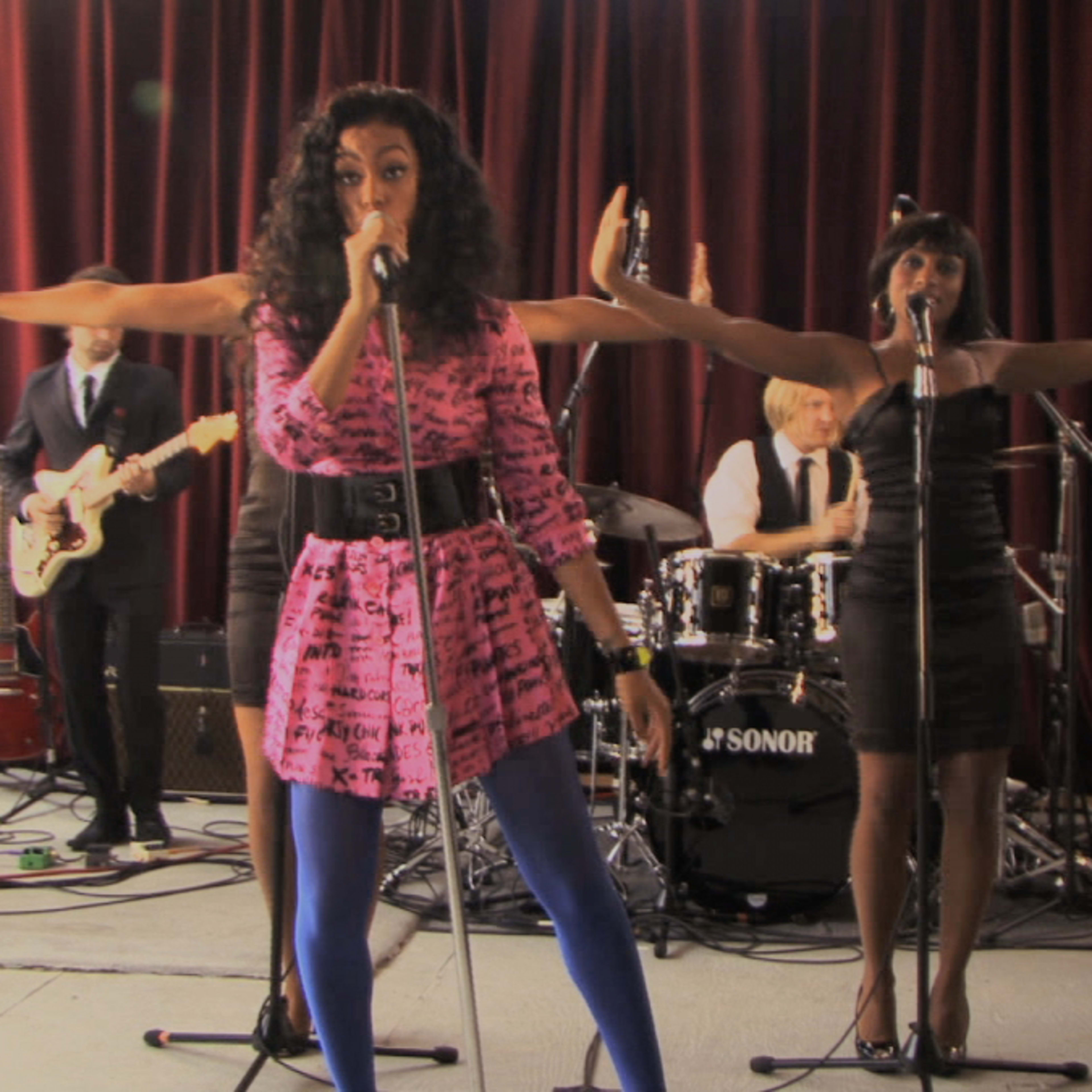 A female singer performs on stage with backup vocalists and a band in a studio setting.