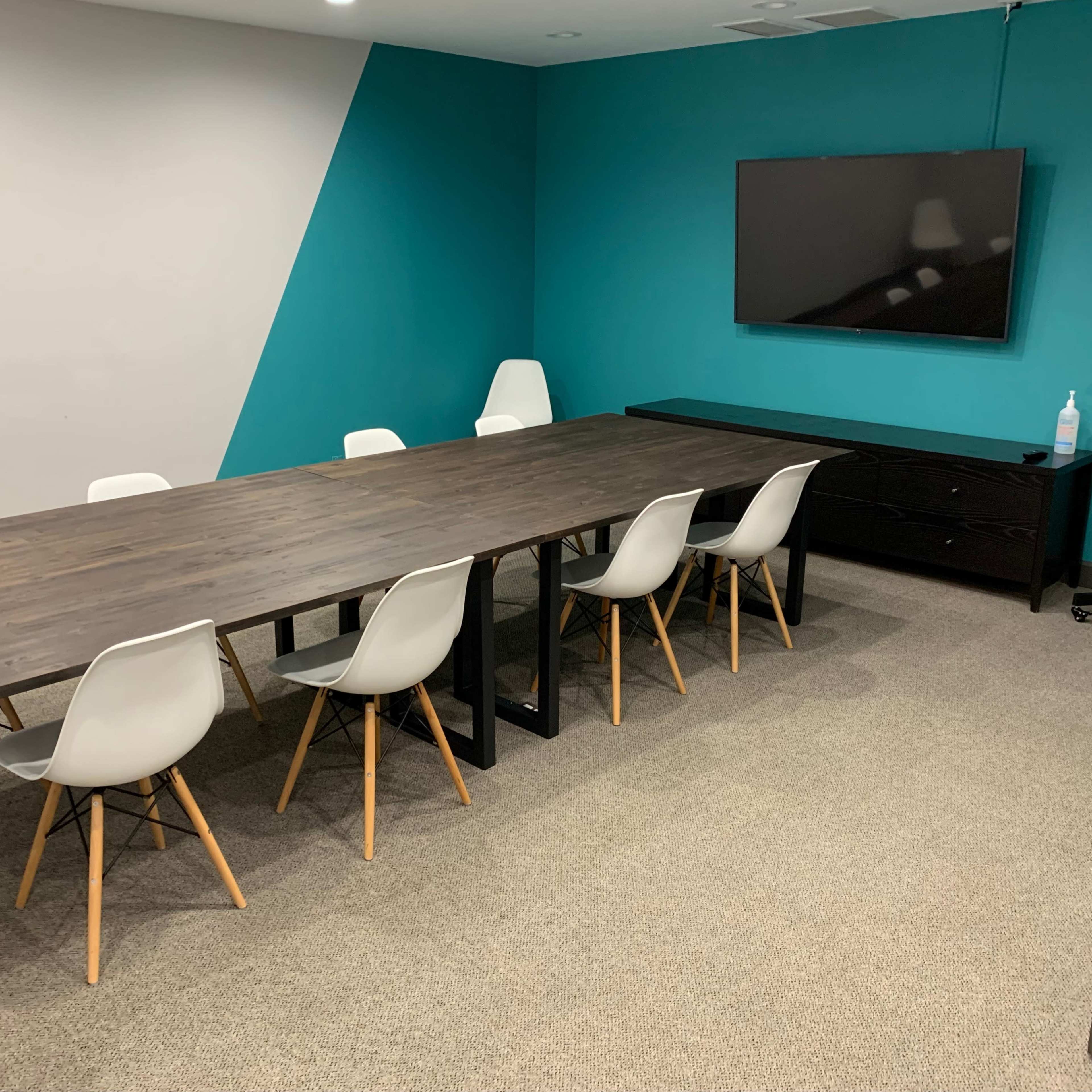 A conference room with a long wooden table surrounded by white chairs, a television mounted on the wall, and a side cabinet.