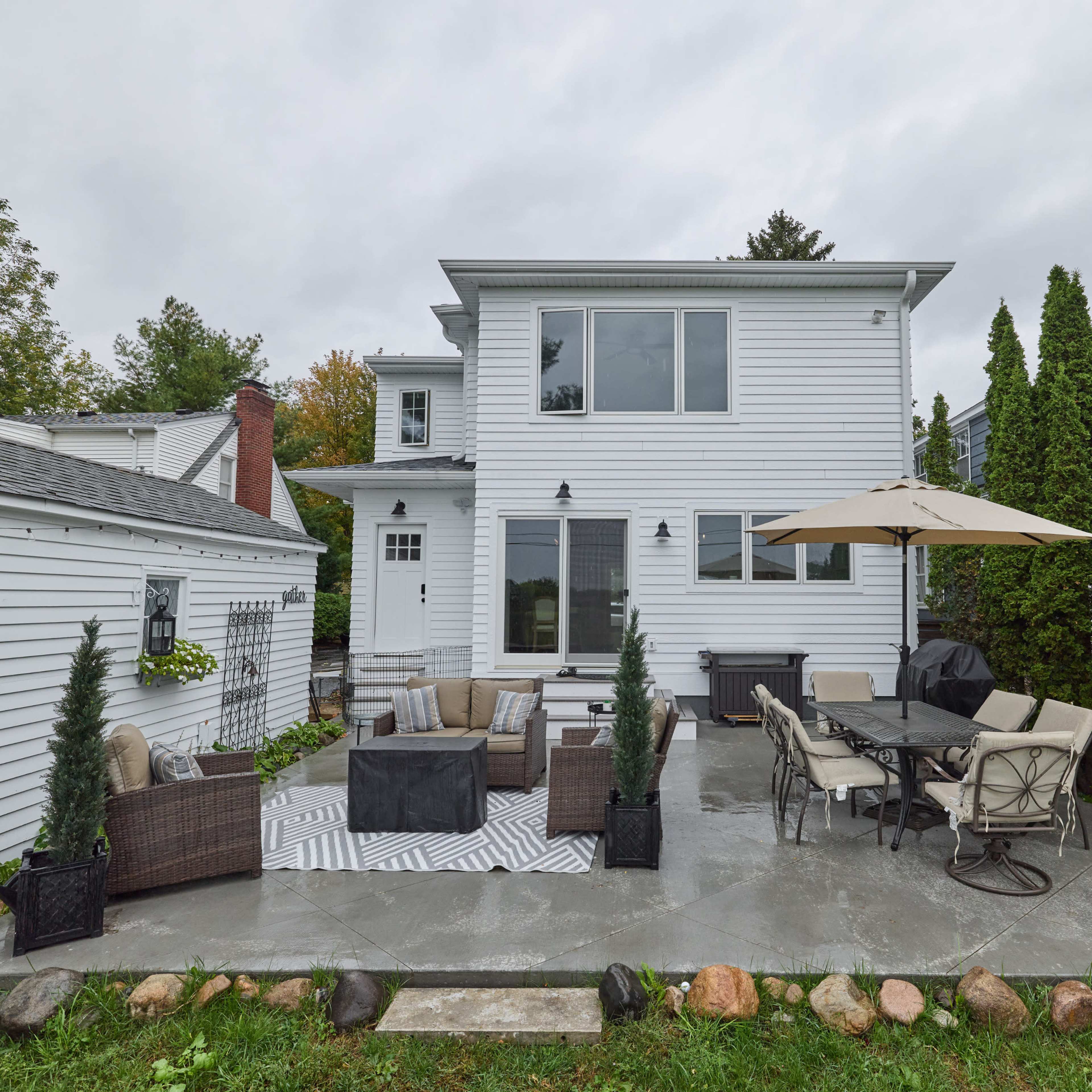 A modern two-story house with a patio featuring outdoor furniture, an umbrella, and tall greenery, situated in a landscaped yard.