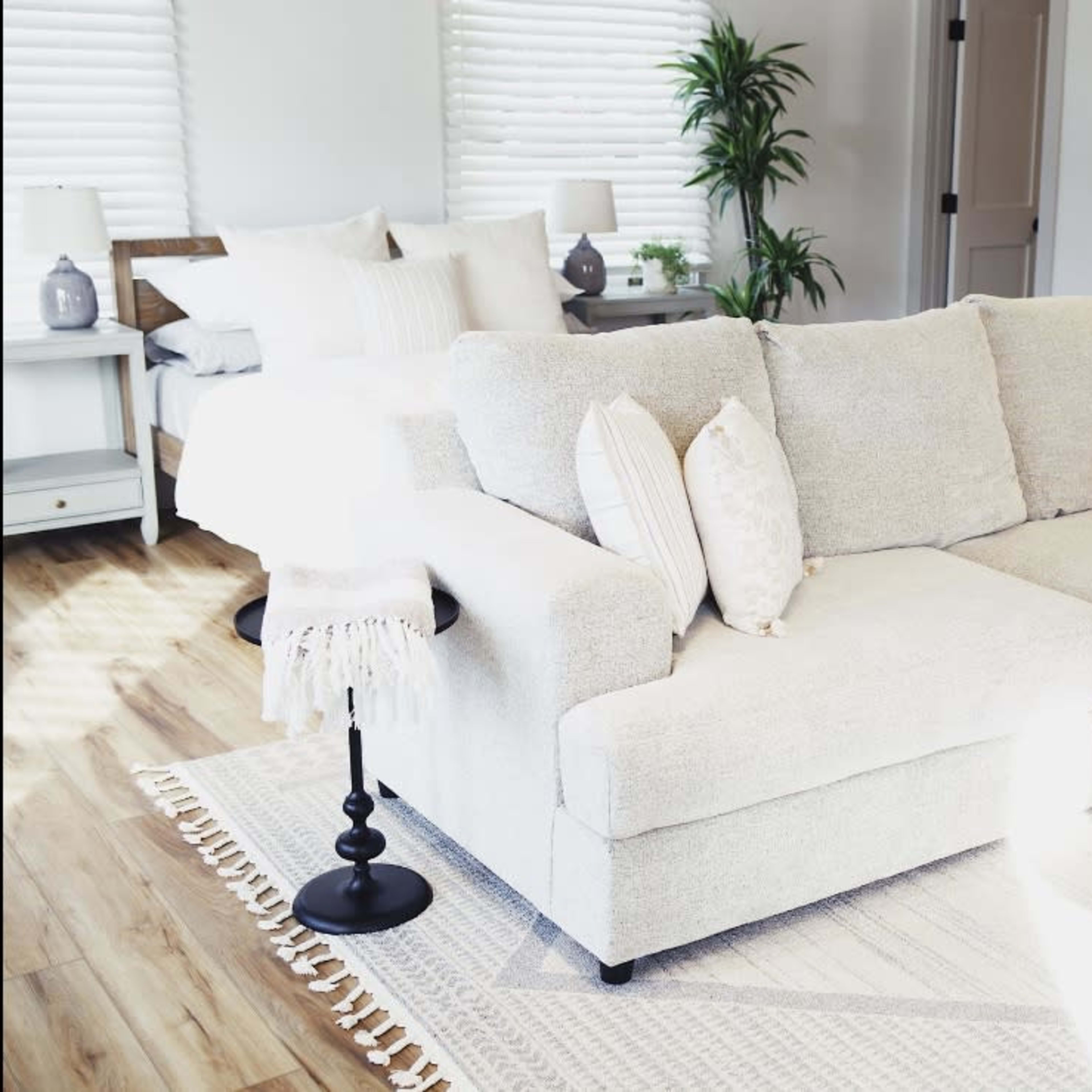 A light-colored sectional sofa is positioned in a cozy room with a bed in the background, accompanied by a wooden coffee table and a small plant.