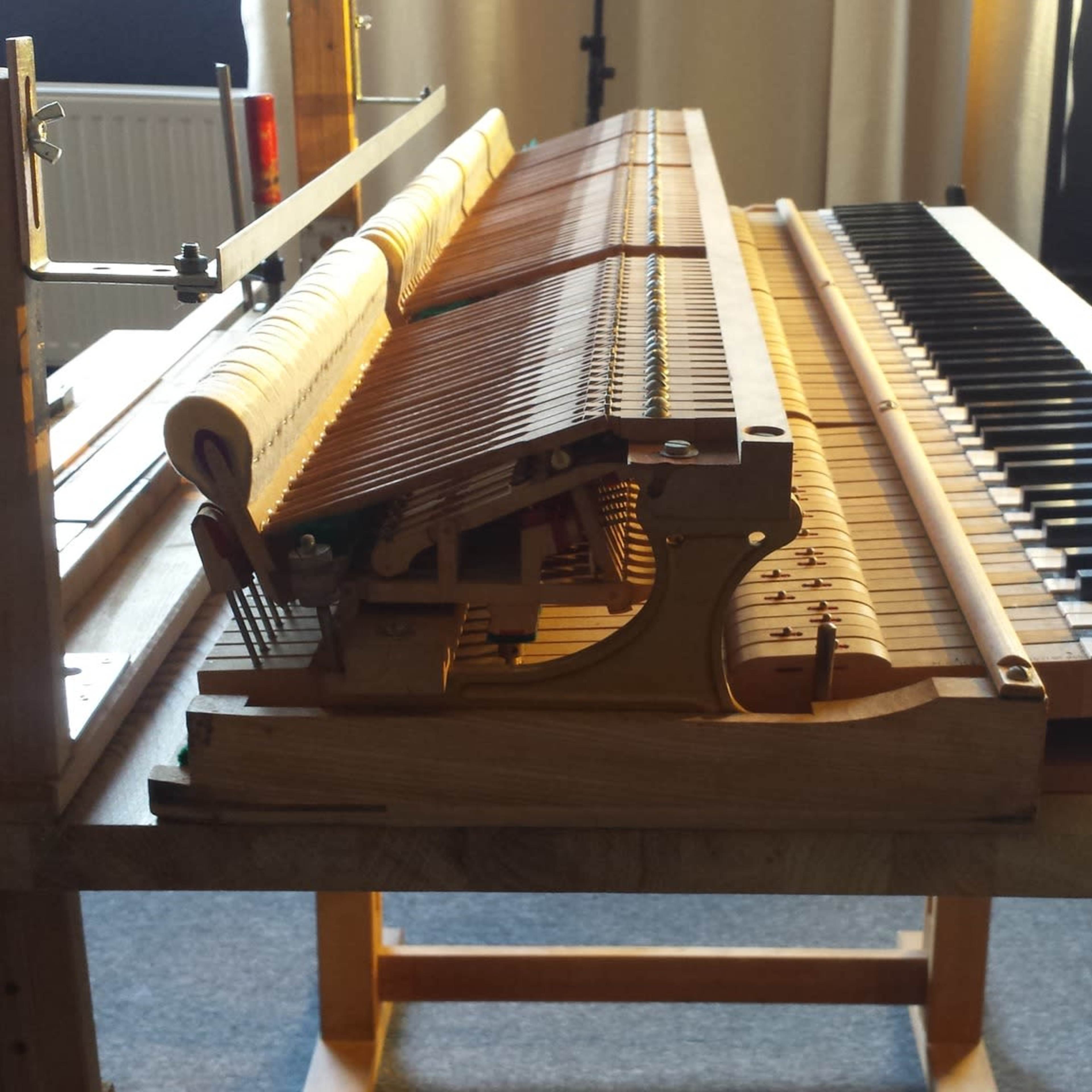 An open grand piano with visible internal mechanisms and keys is positioned on a wooden stand in a well-lit room.