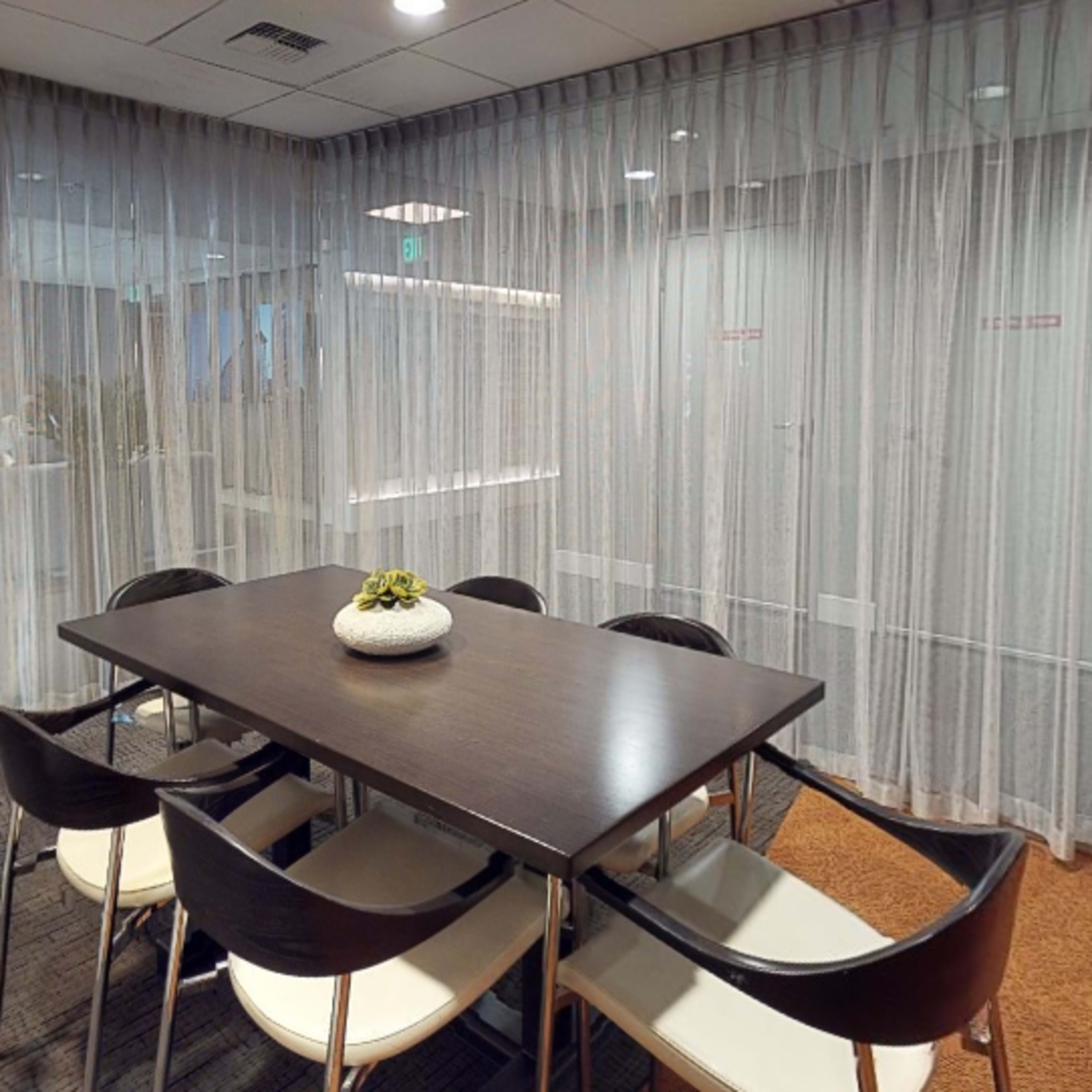 A conference room with a dark wooden table surrounded by white chairs, enclosed by sheer curtain panels.