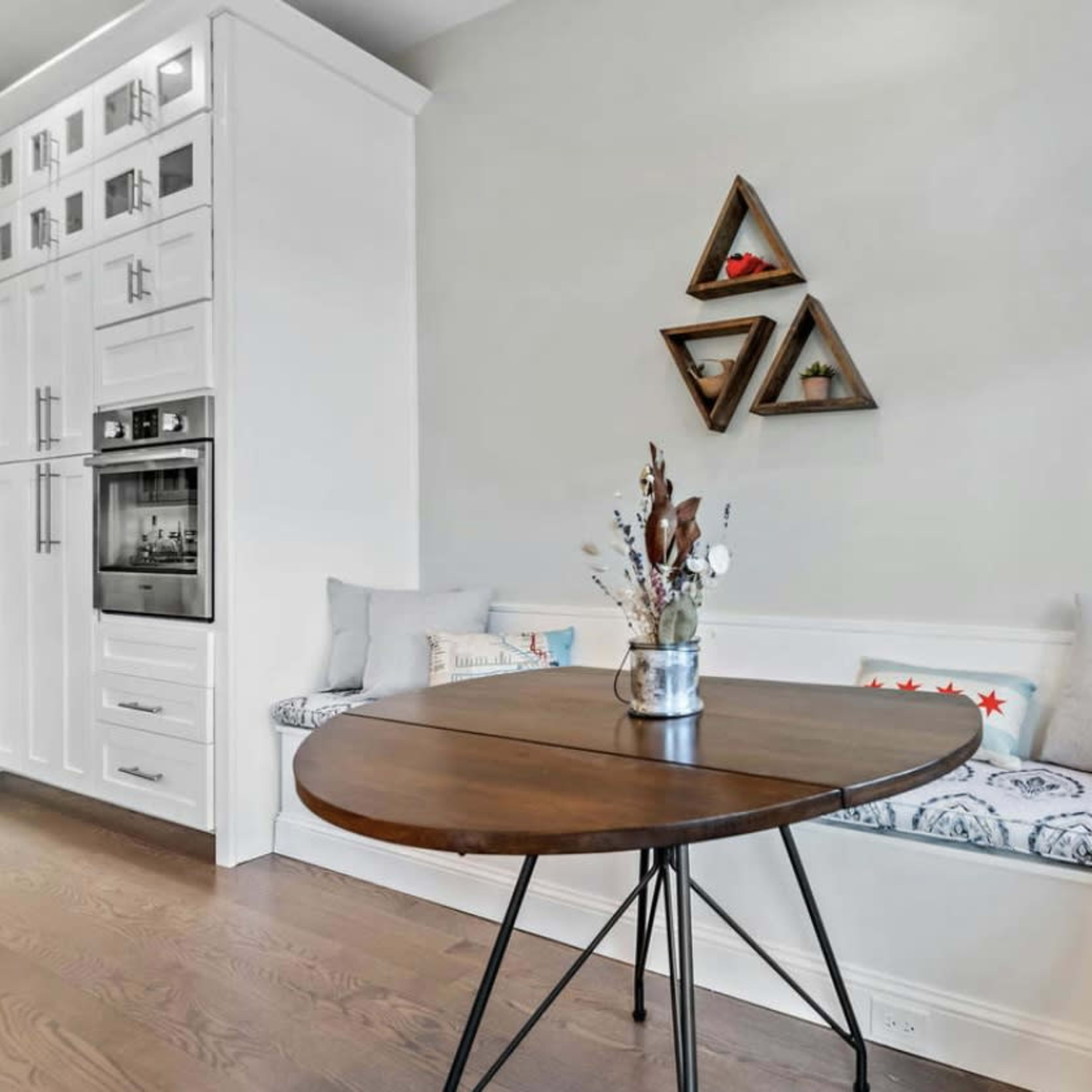 A round wooden table sits in front of a built-in bench with decorative pillows against a light-colored wall featuring triangular wall art.