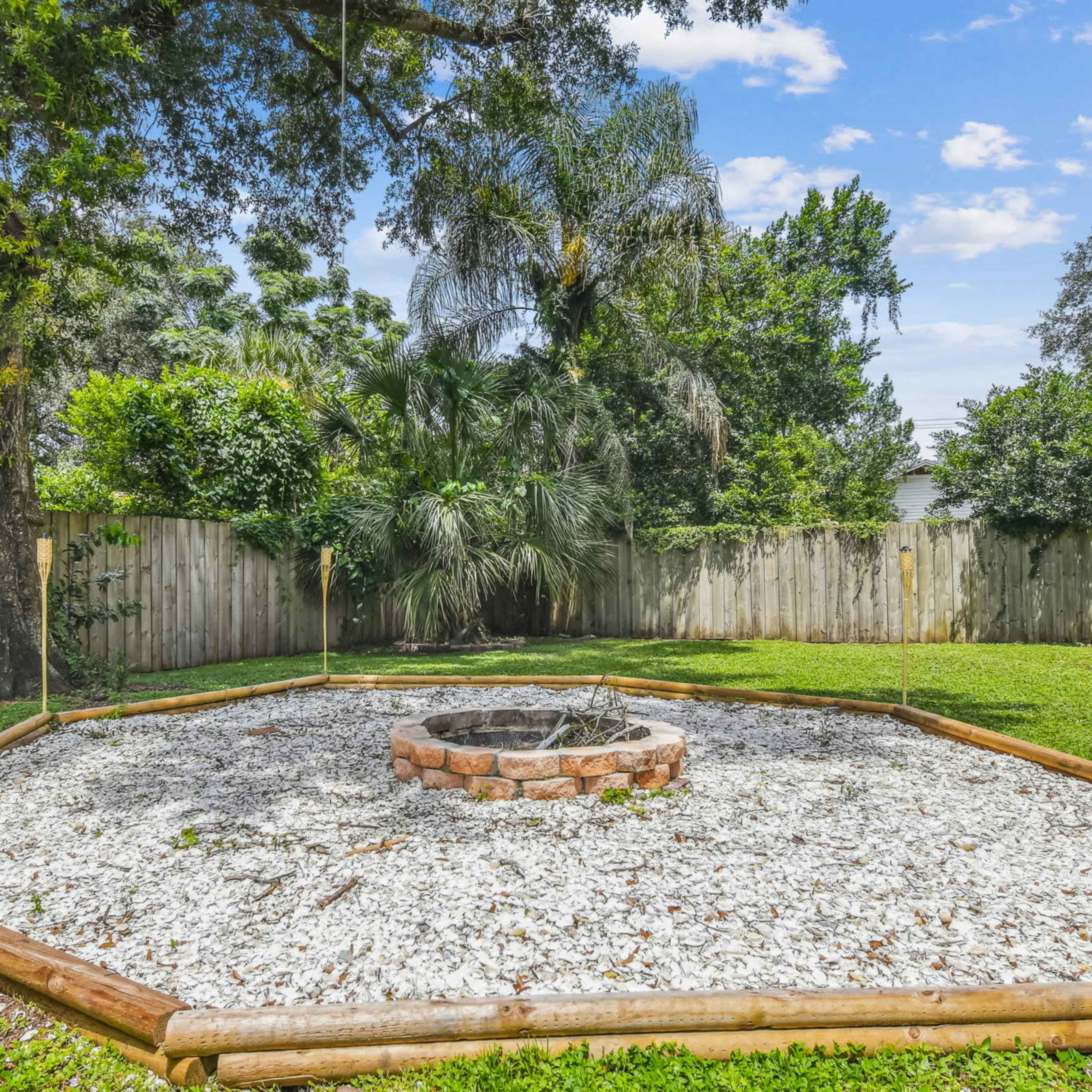 A circular fire pit surrounded by gravel and wooden edging is located in a well-maintained yard, with tiki torches placed around its perimeter.