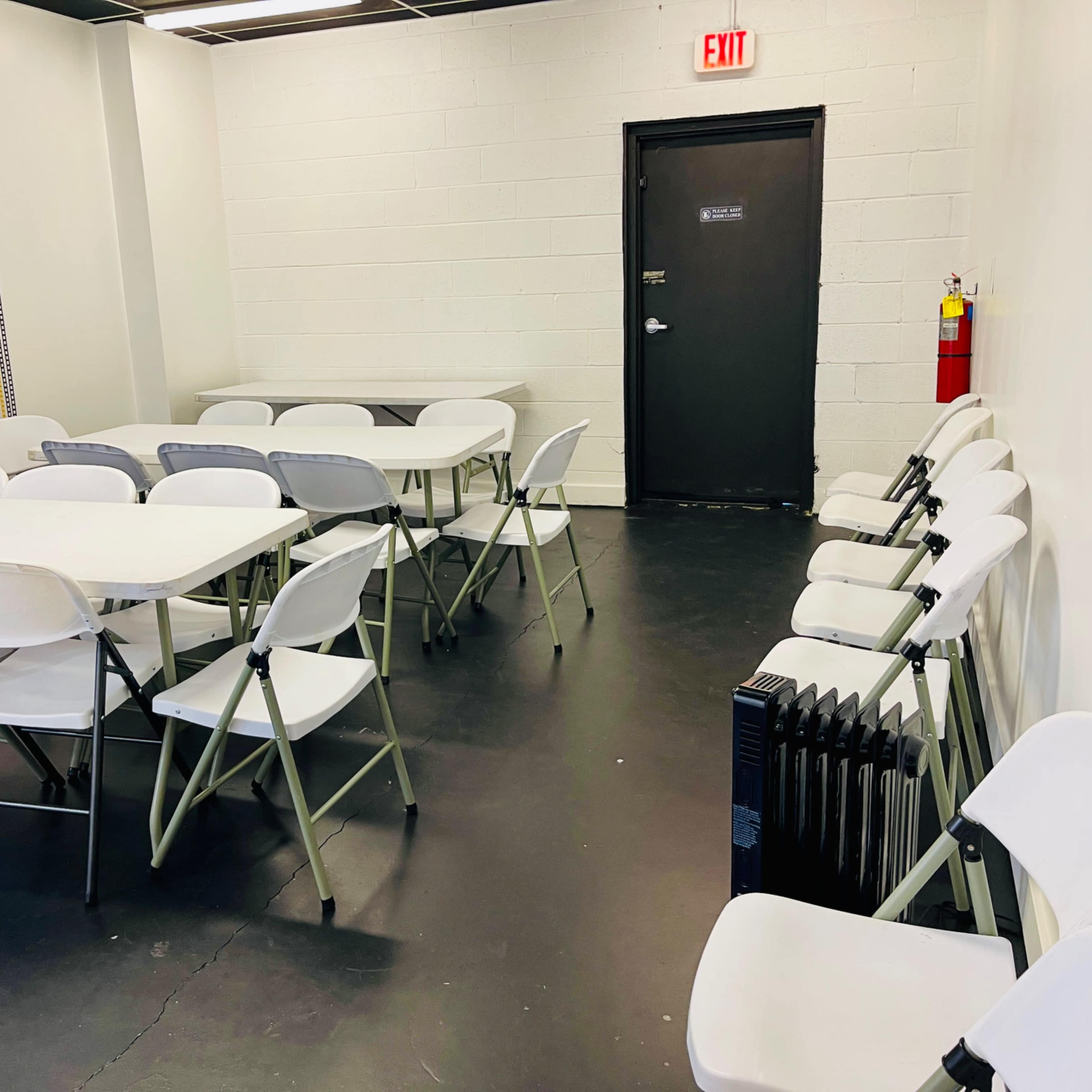 A room with several white folding tables and chairs arranged around, a black door, and a radiator along the wall.