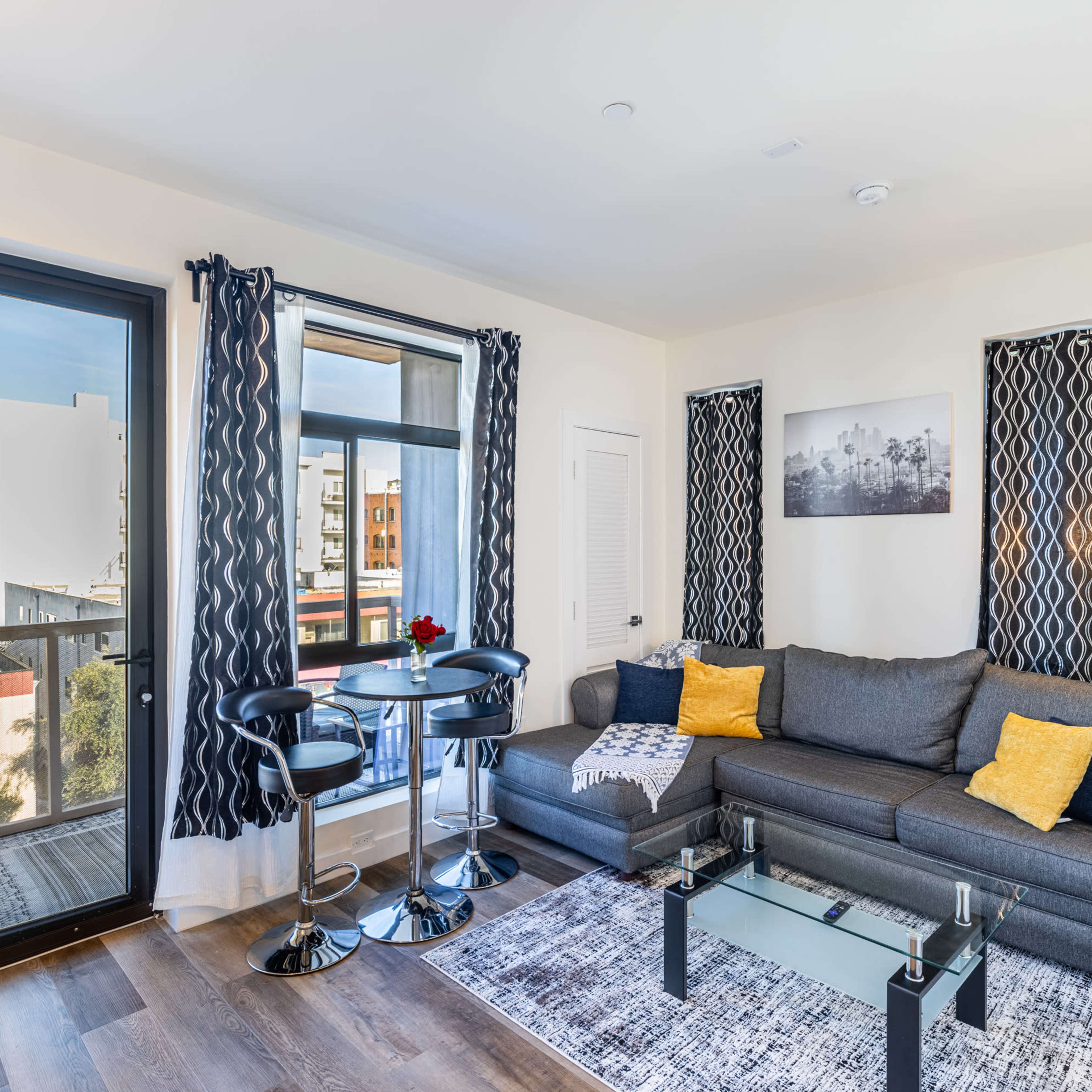 A cozy living room features a gray couch, a glass coffee table, and a small dining area with two chairs, complemented by large windows and modern decor.