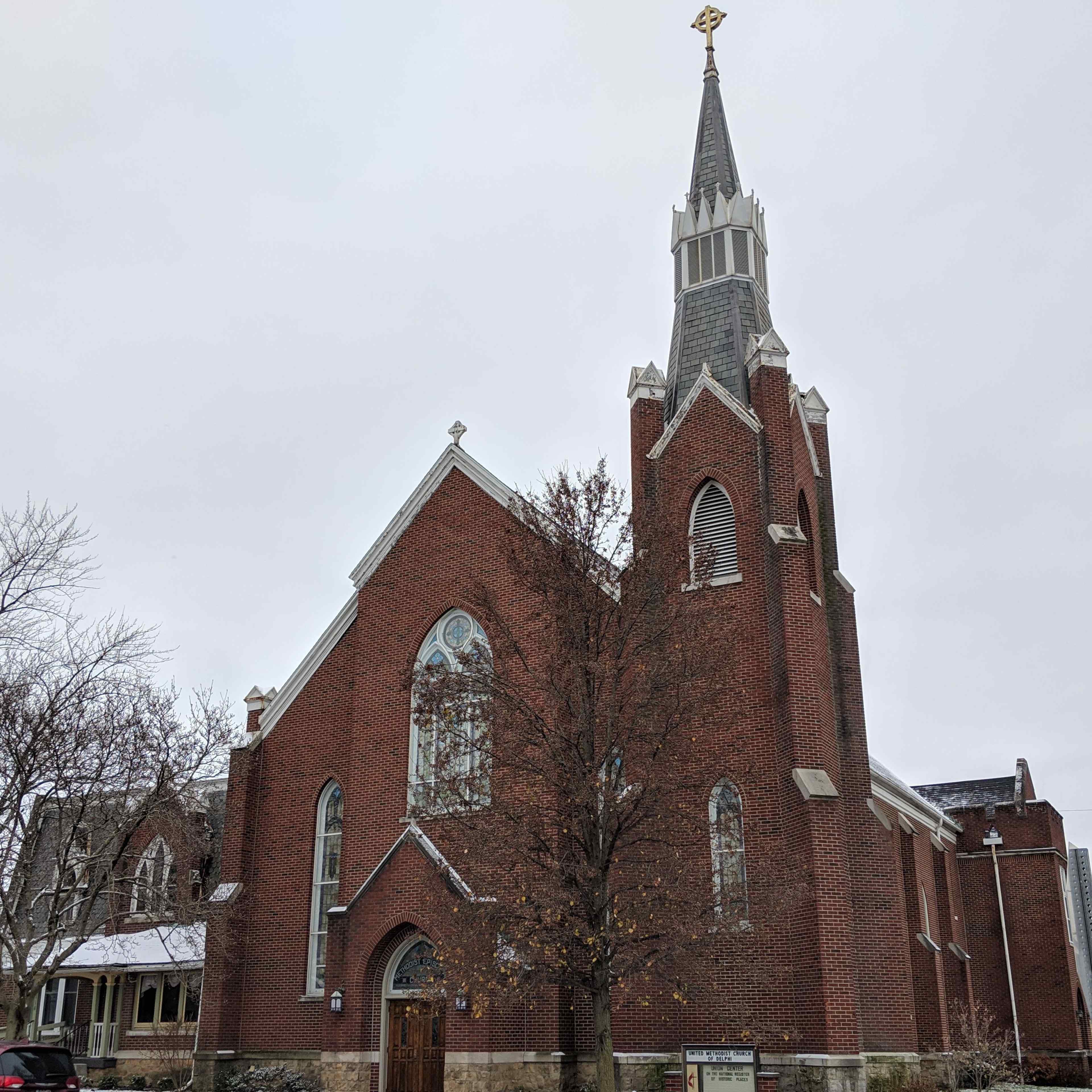 A brick church with a tall steeple and a large arched window stands beside a bare tree in a cloudy sky.