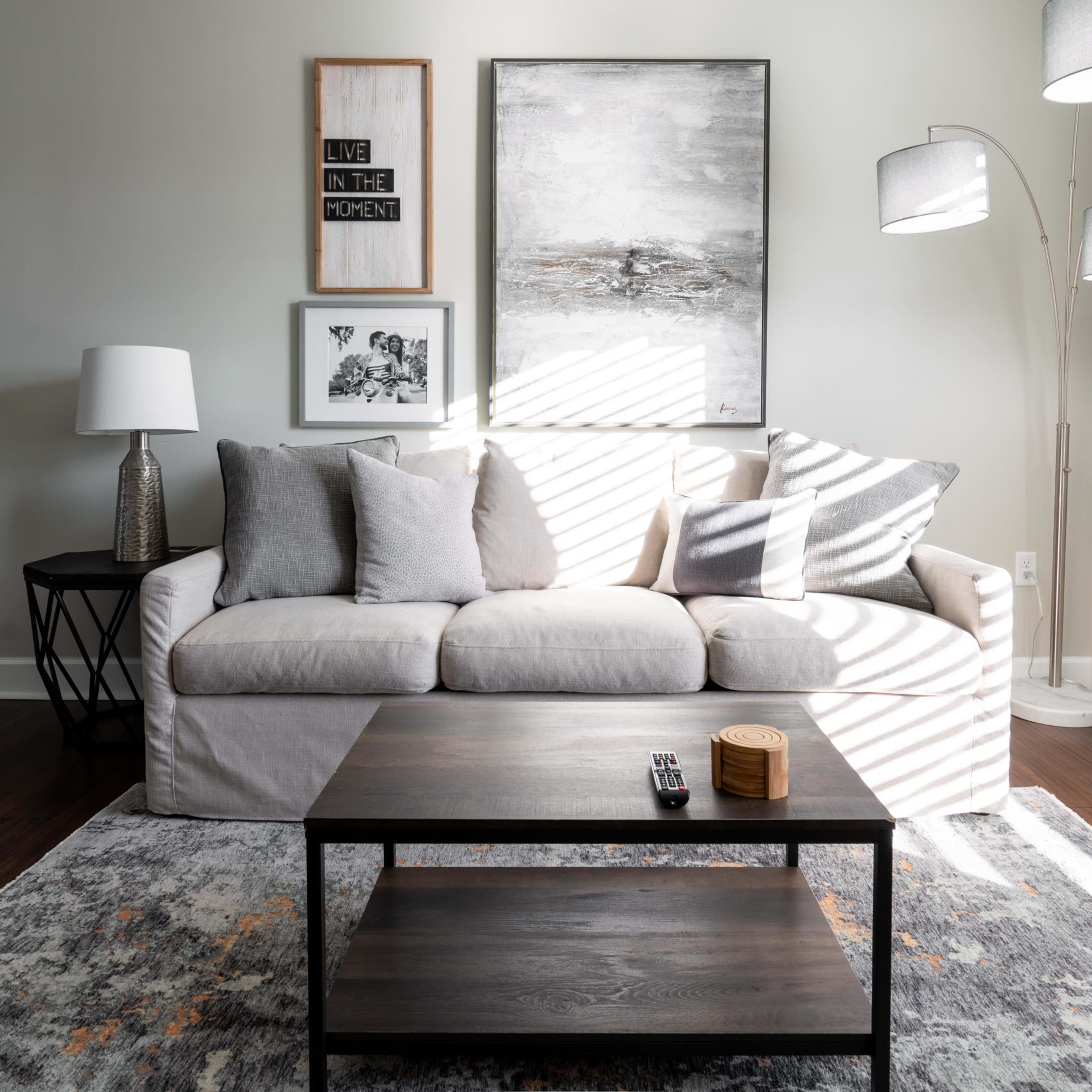 A living room features a light-colored sofa with several cushions, a dark wood coffee table, and a tall floor lamp, illuminated by sunlight casting striped shadows.